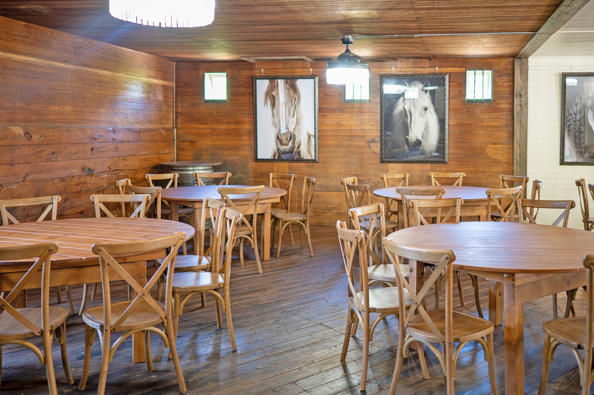 A rustic, wood-paneled room features several round wooden tables surrounded by wooden chairs, with framed horse art.
