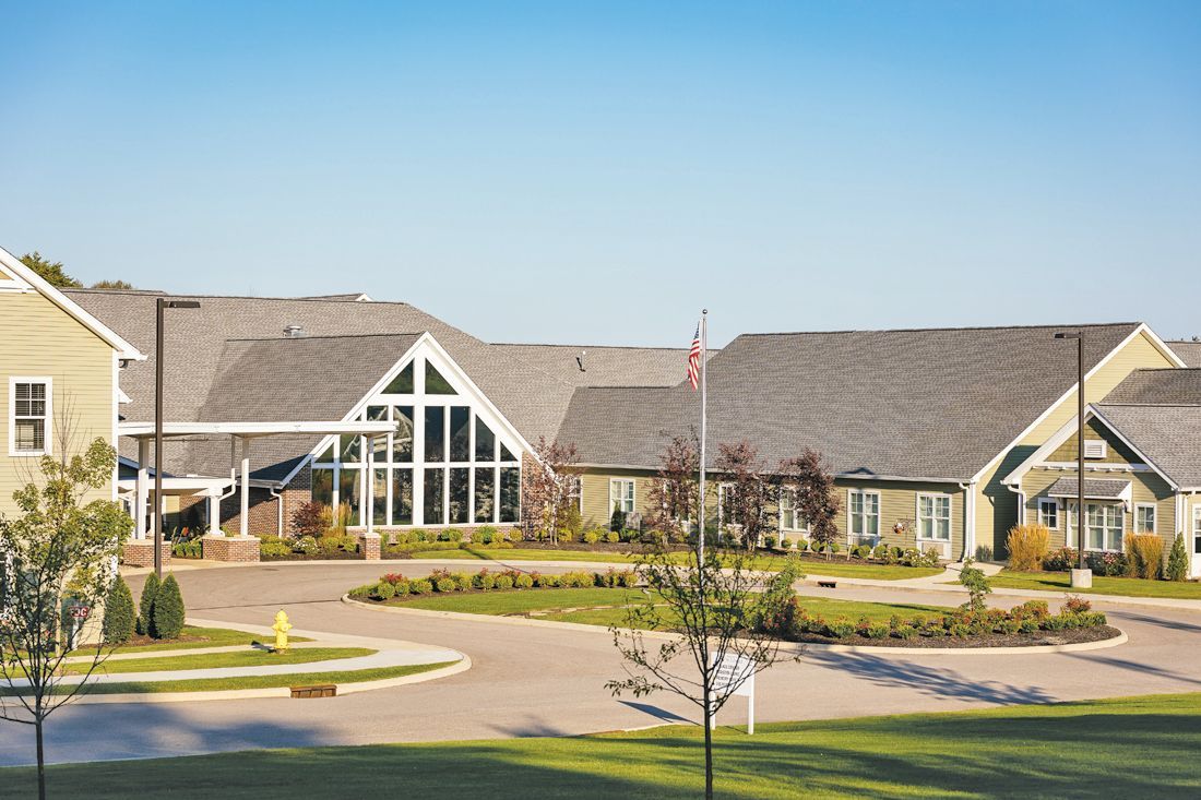 A one-story assisted living facility with light green siding, a grey shingled roof, and a large triangular window.