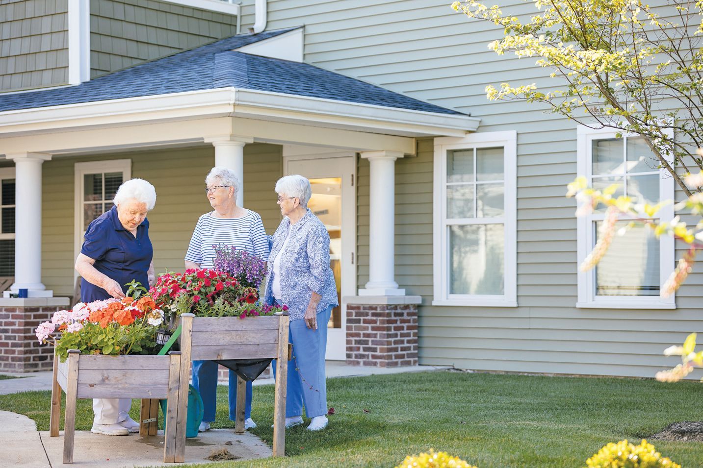Three people tend to a wooden garden planter outside a building with light green siding and white columns.