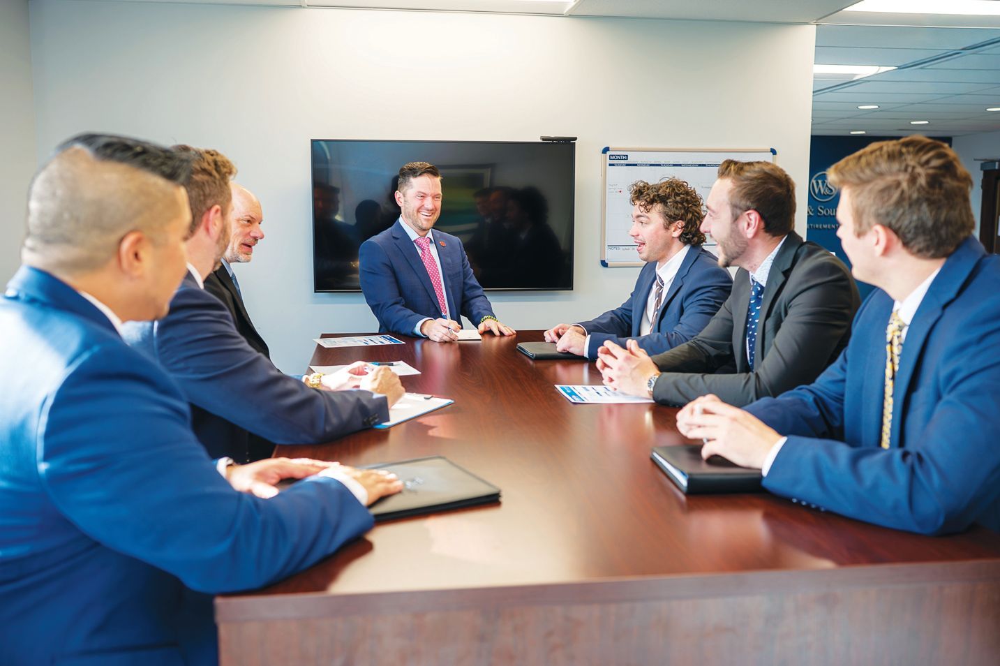 A group of professionals in suits sitting around a conference table in an office, discussing business.