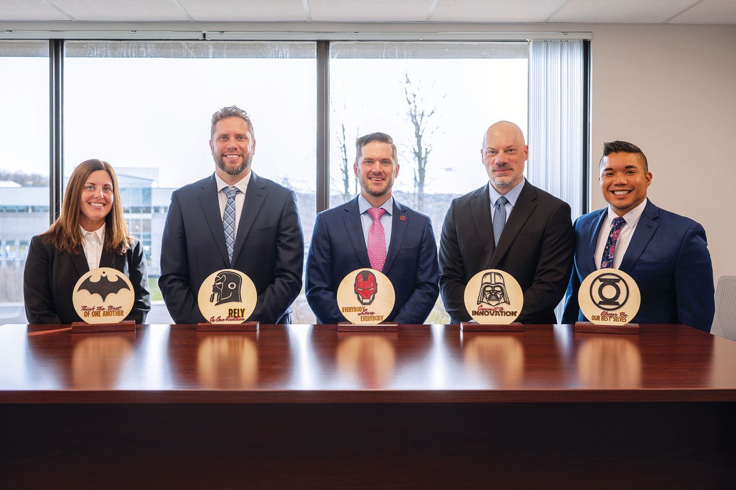 Five people in business suits stand behind a conference table, each holding a circular superhero-themed plaque.
