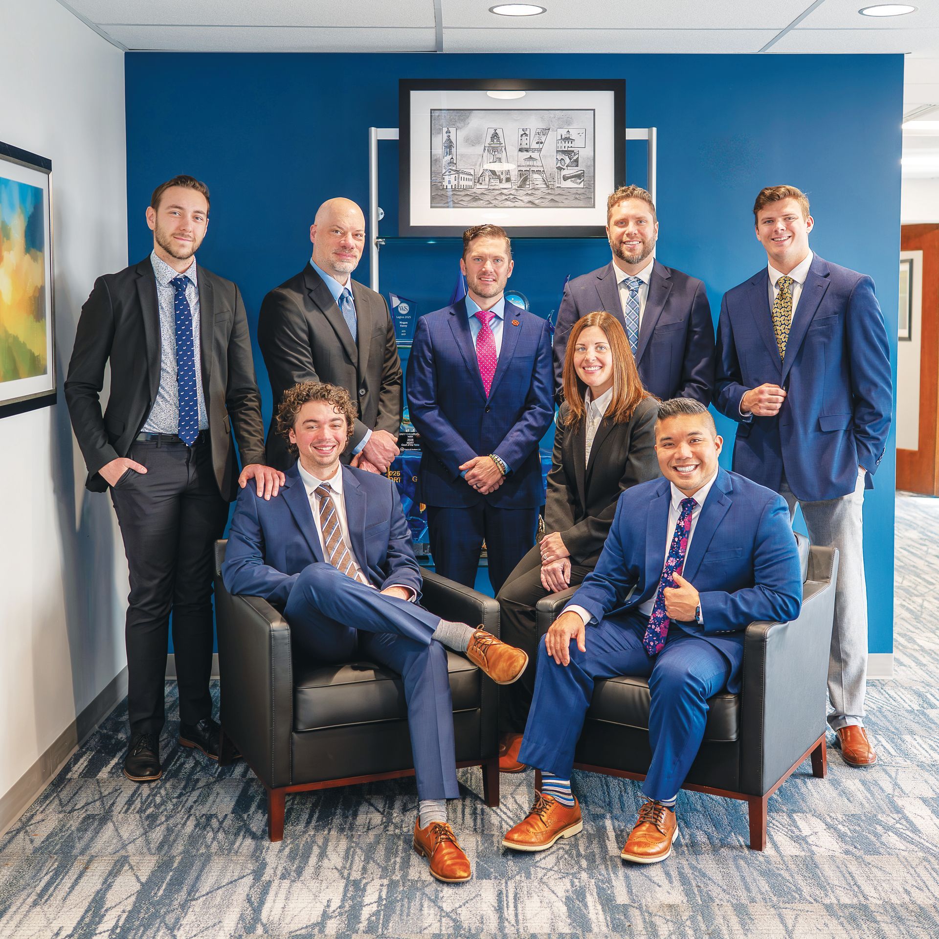 A group of eight professionals in business attire posed in an office, two seated in chairs and six standing behind them.