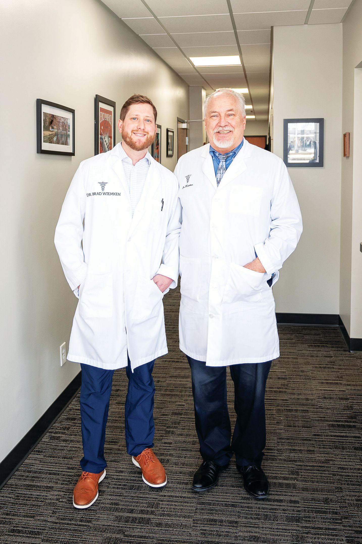 Two professionals in white lab coats standing in a hallway with a patterned carpet, smiling toward the camera.