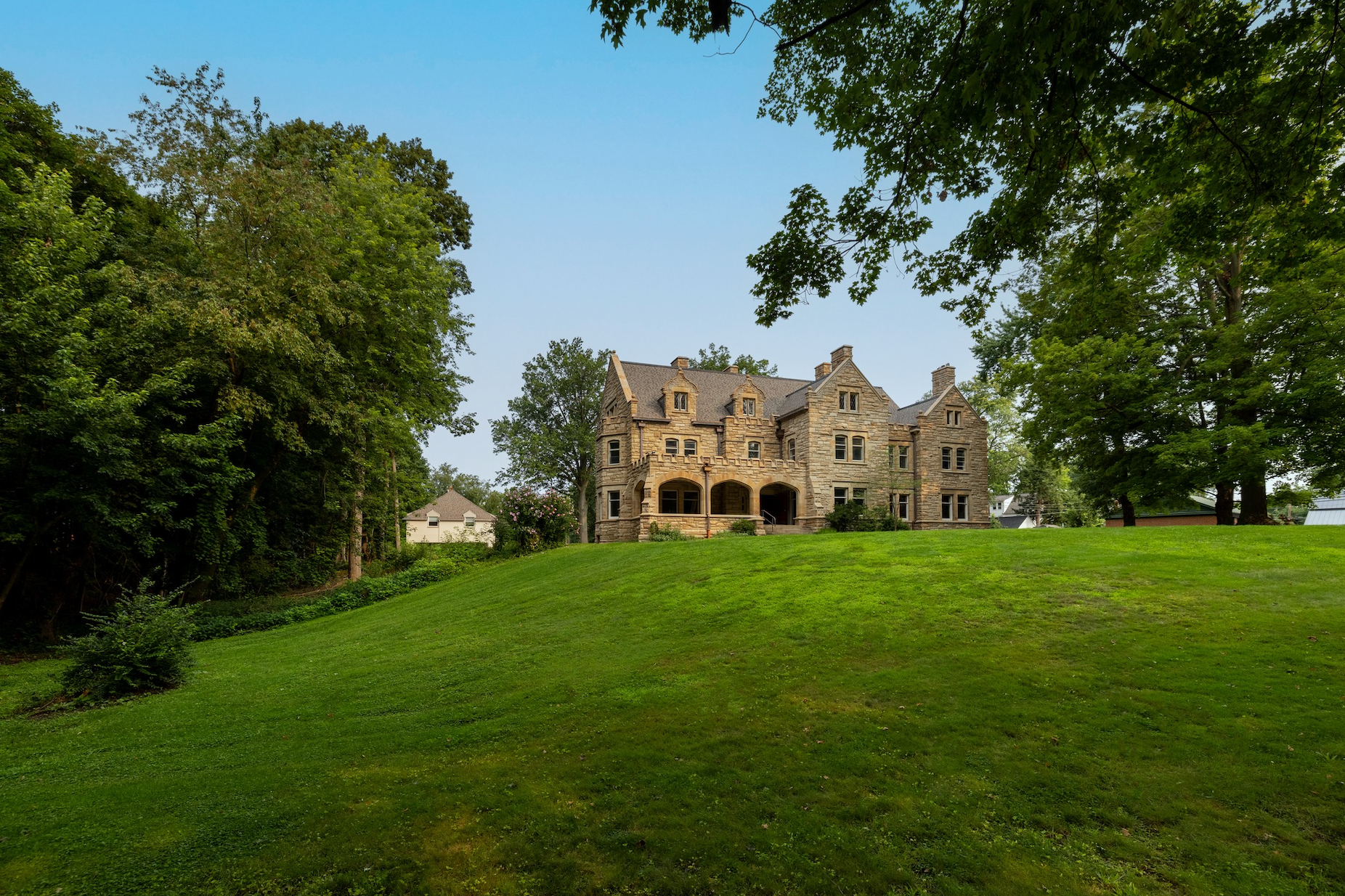 A large stone mansion with multiple gables sits atop a grassy hill surrounded by mature green trees under a blue sky.