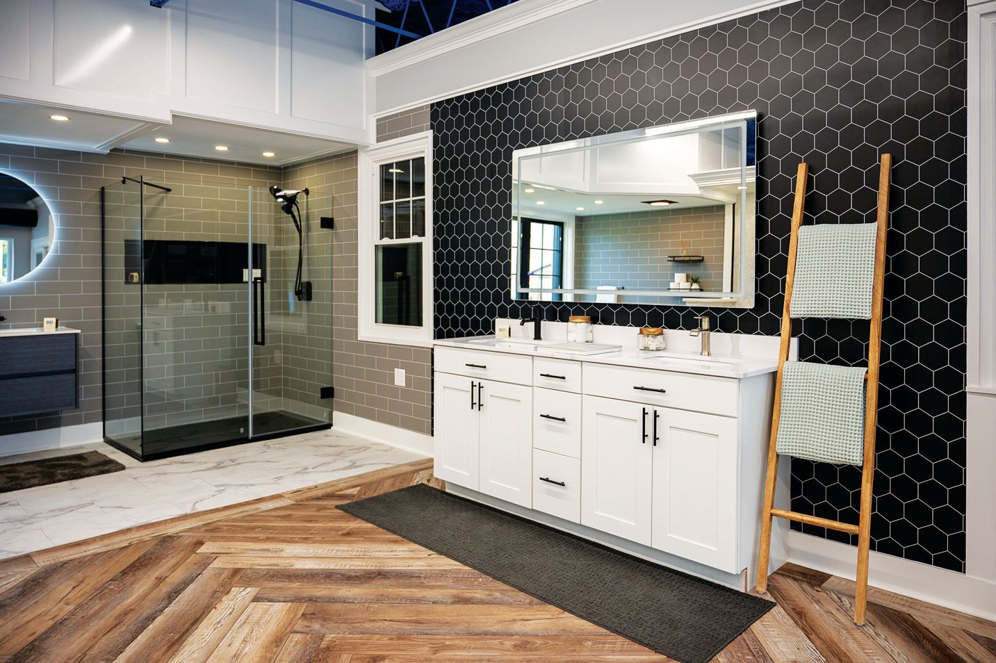A modern bathroom with a glass-enclosed shower, white vanity, black hexagonal tile wall, and wood-patterned floors.