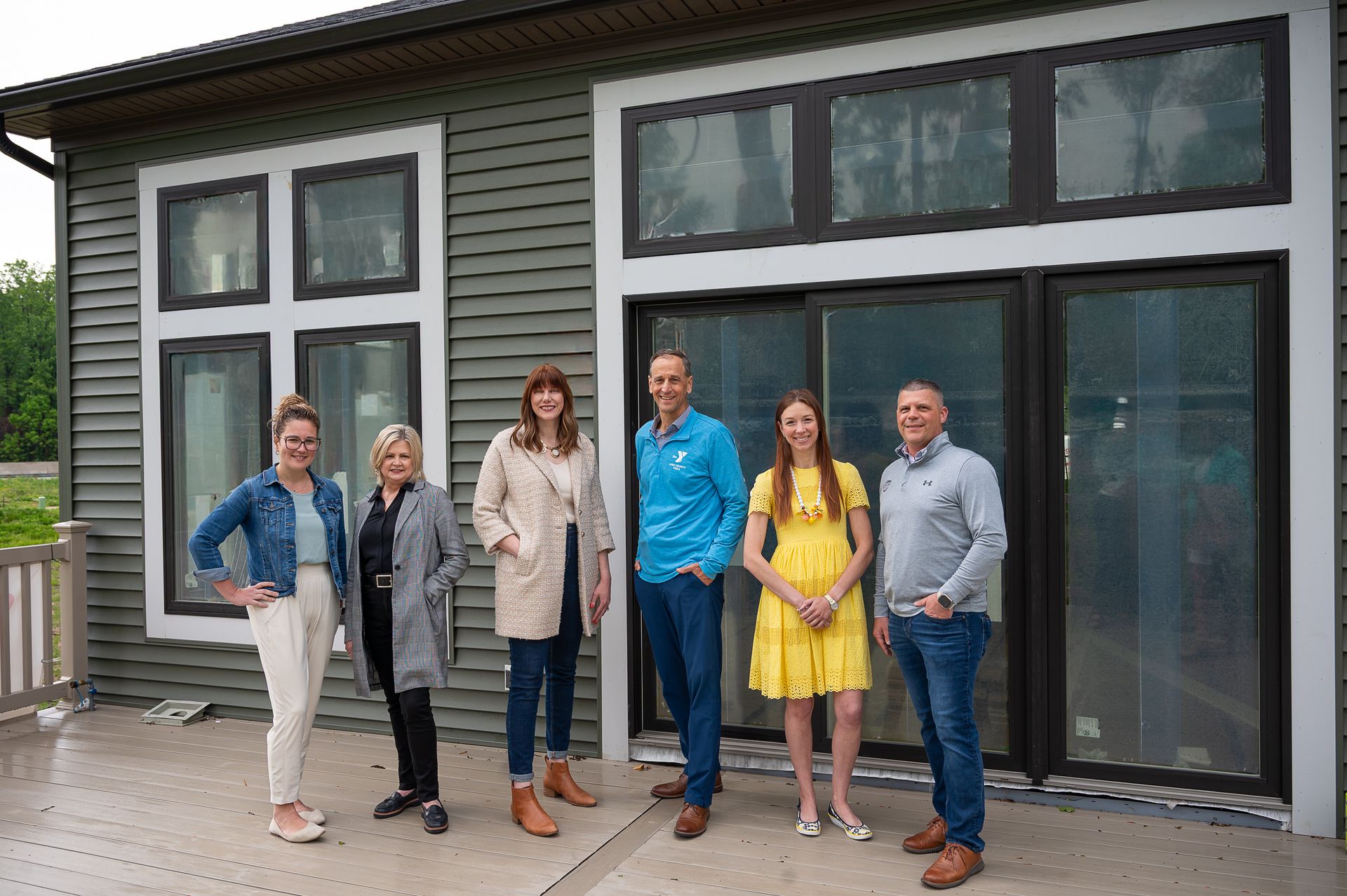 Six people stand in a line on a deck in front of a modern house with grey siding and large windows.