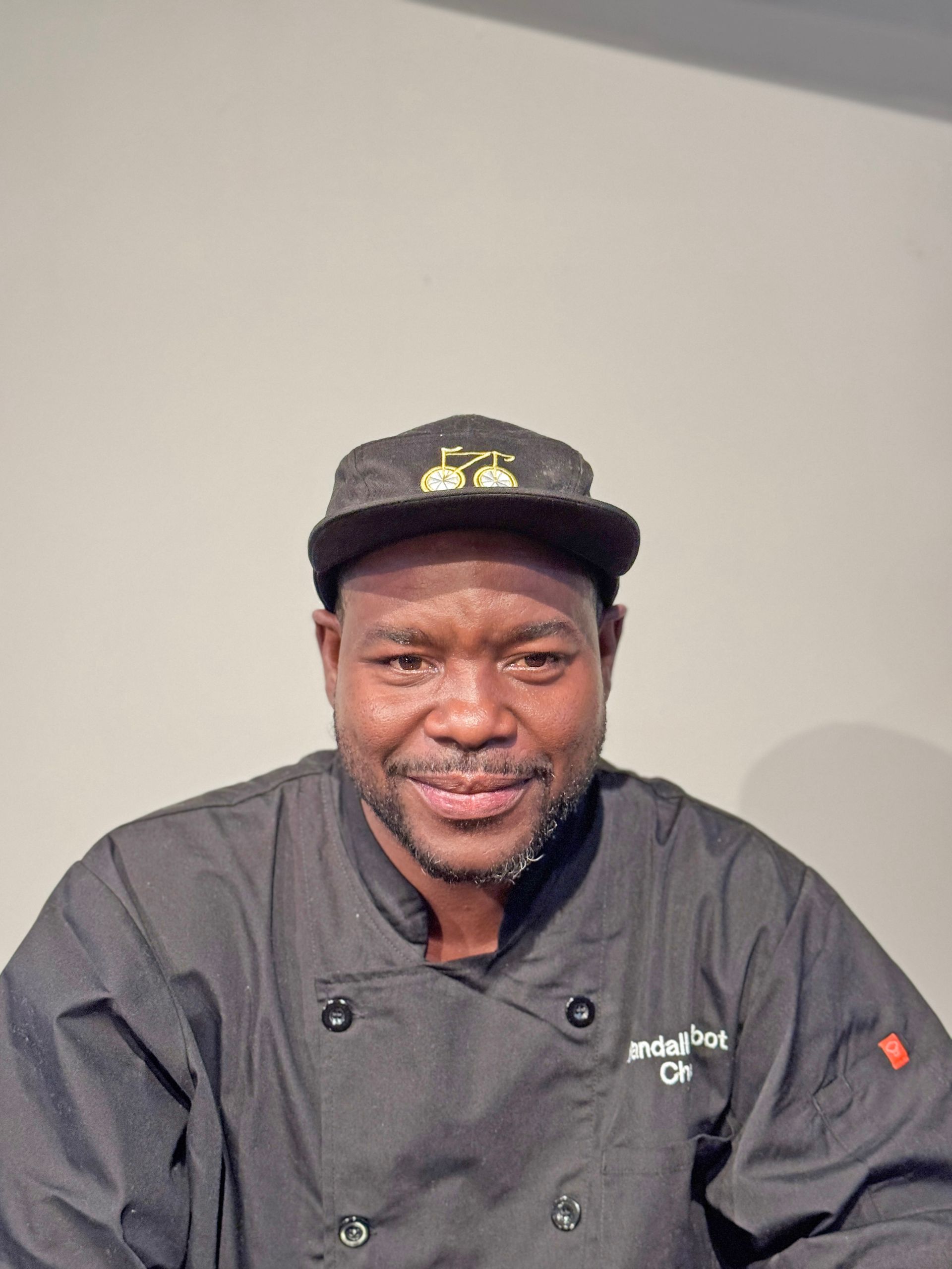 A person wearing a black chef’s coat and a black baseball cap with a gold emblem smiles against a plain, light background.