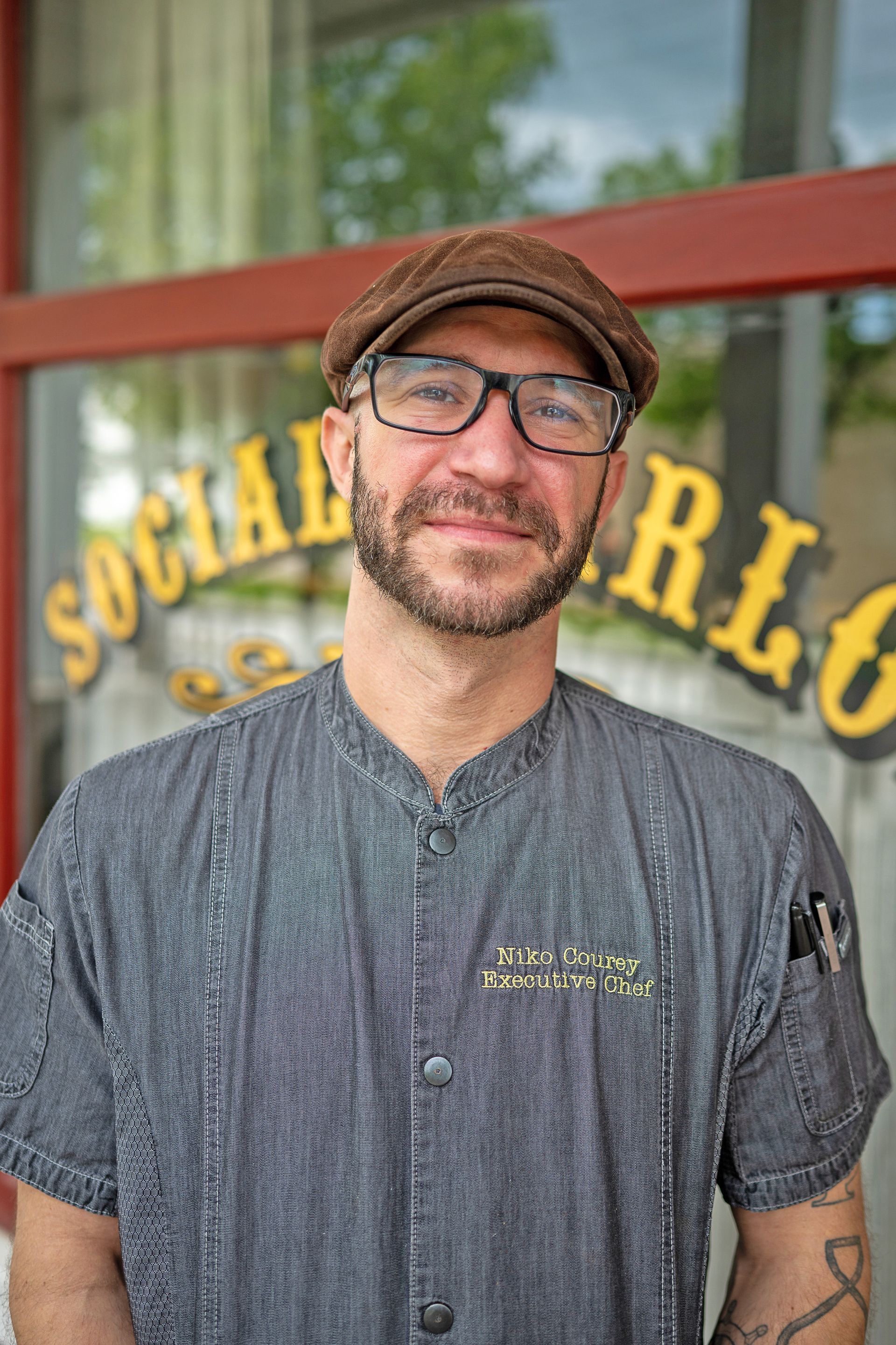 A person in a black chef’s coat leaning against a wooden pillar in a restaurant dining area.