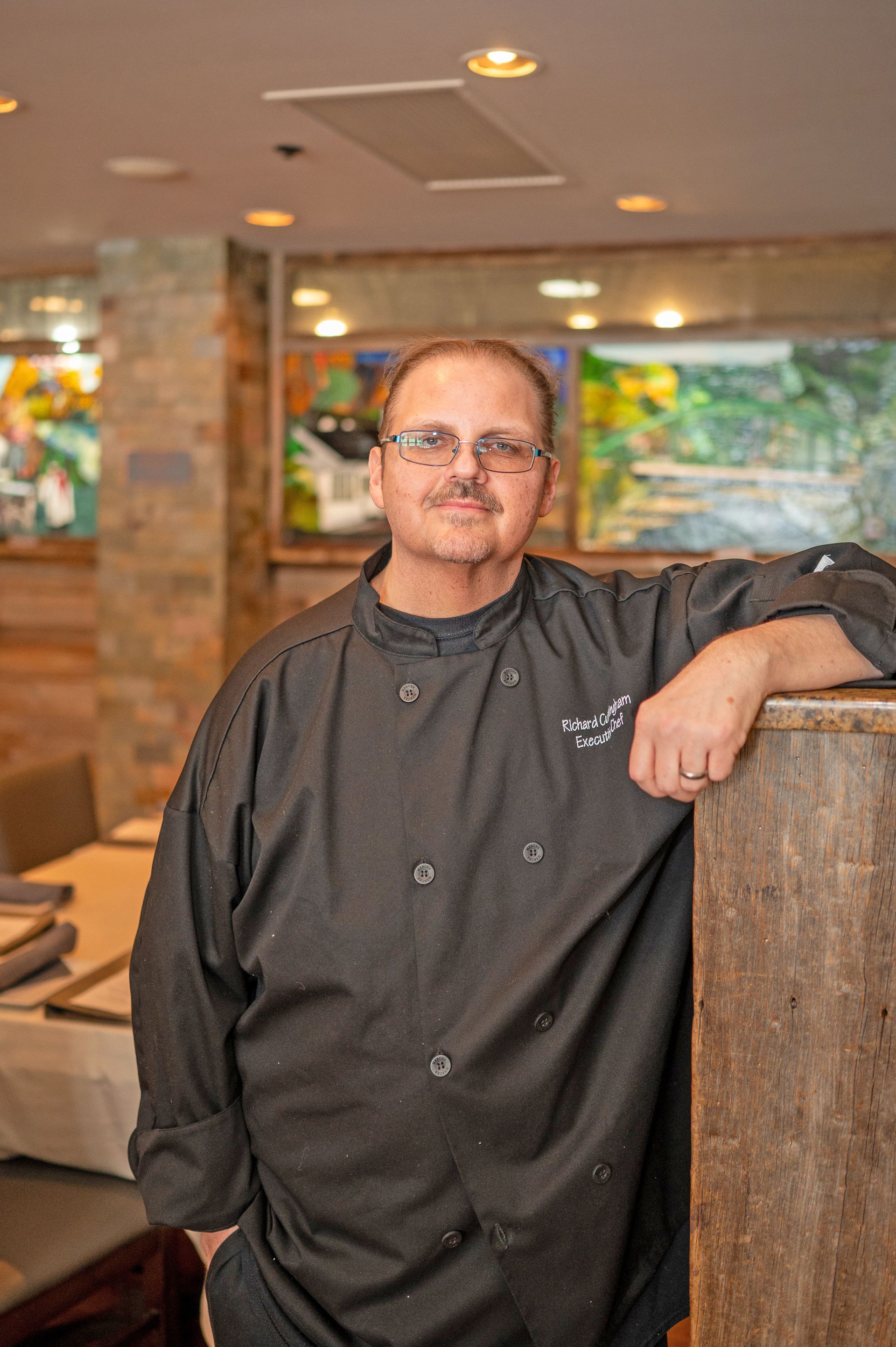 A person in a black chef’s coat leaning against a wooden pillar in a restaurant dining area.