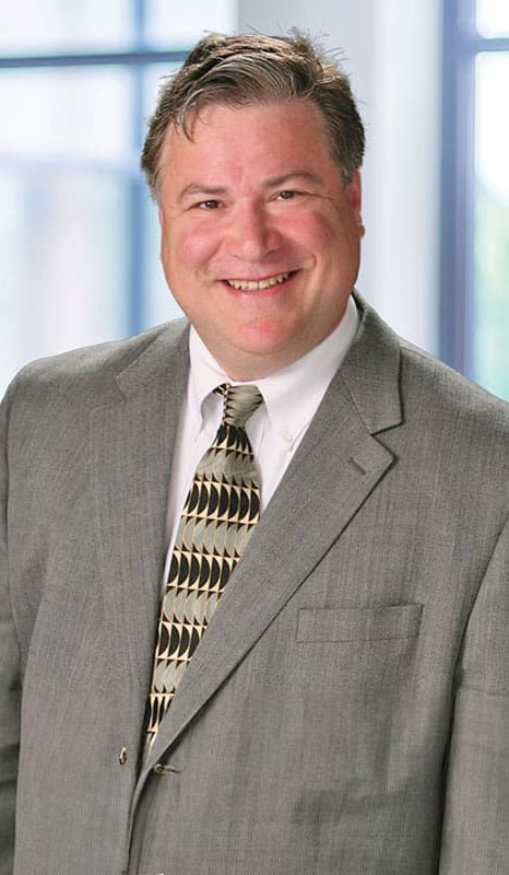A smiling person in a grey suit and a patterned tie, posing against a blurred indoor backdrop.