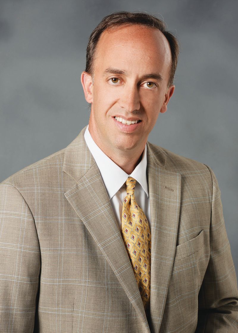 A professional headshot of a person in a tan blazer, white shirt, and yellow patterned tie against a gray background.