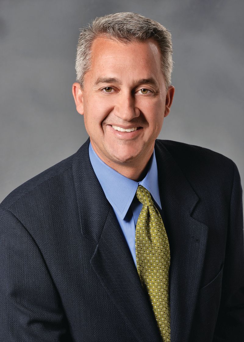 A professional portrait of a smiling person wearing a navy blazer, light blue shirt, and a textured olive-green tie.