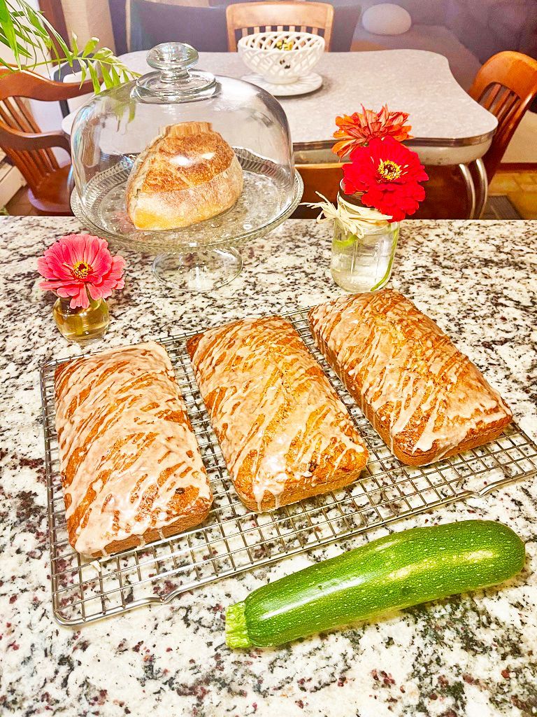 Three glazed loaves of zucchini bread on a cooling rack next to a fresh zucchini, with more bread under a glass dome.