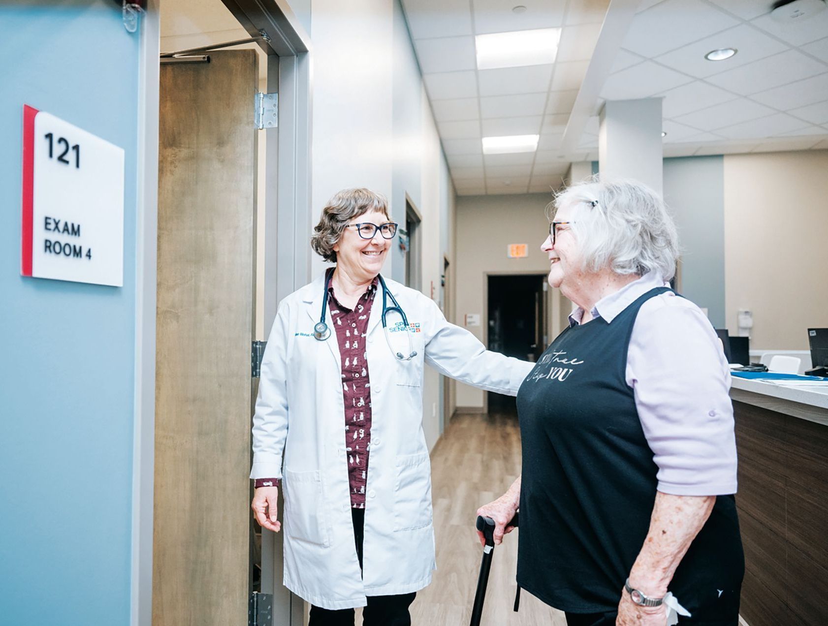 A medical professional in a white coat with a stethoscope smiles at a patient holding a cane in a clinic hallway.