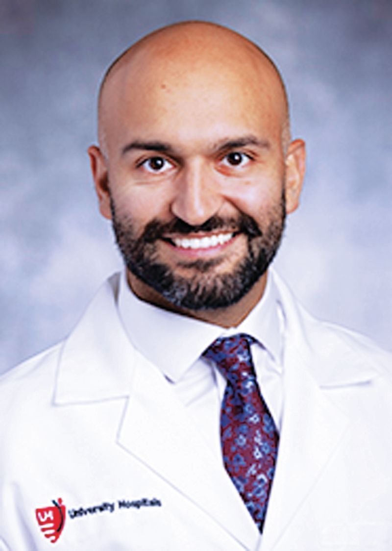A smiling professional wearing a white coat, collared shirt, and patterned tie against a mottled blue background.