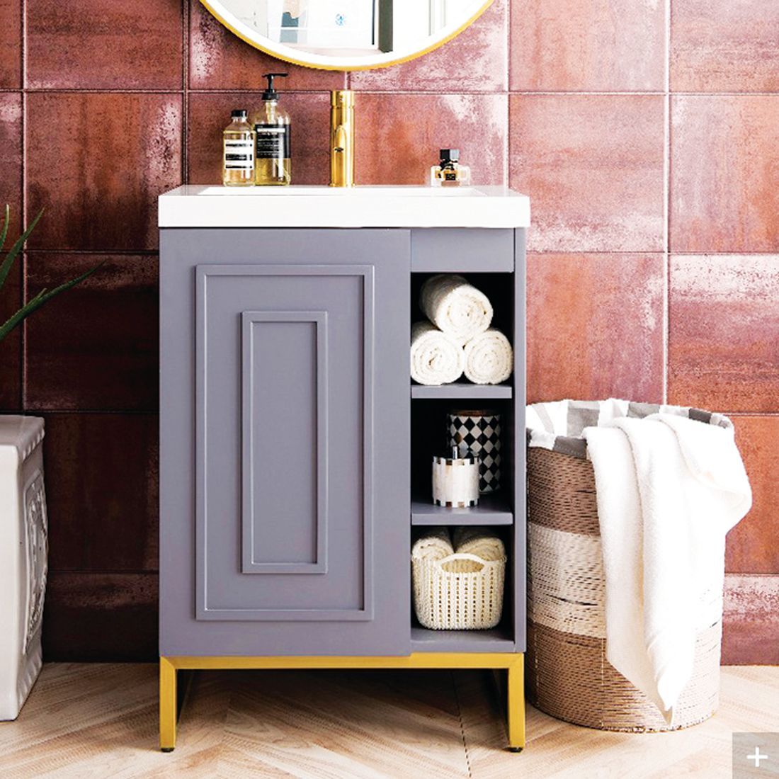 A grey bathroom vanity with gold legs and open shelving next to a woven laundry basket against a rust-colored tile wall.