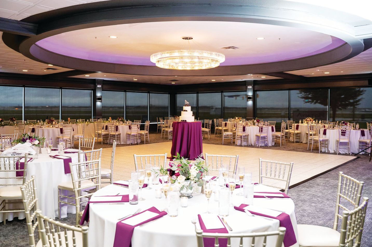 A formal event space with round tables, white linens, purple accents, and a tiered cake on a pedestal in the center.