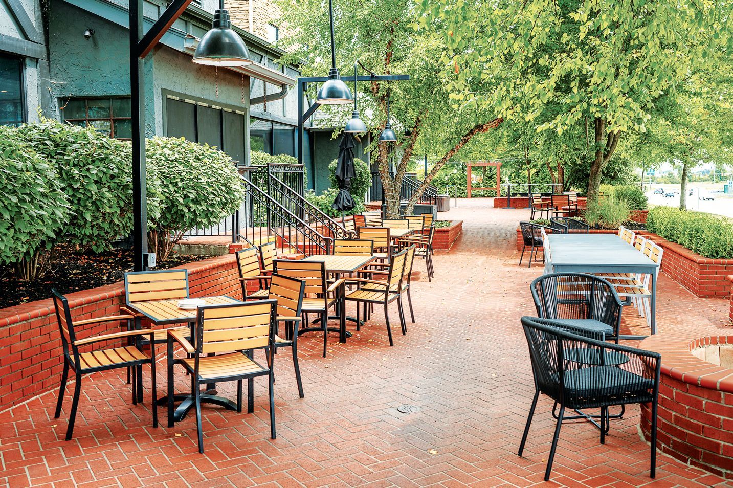 Outdoor restaurant patio with wood and metal tables and chairs on a red brick floor surrounded by green trees and bushes.