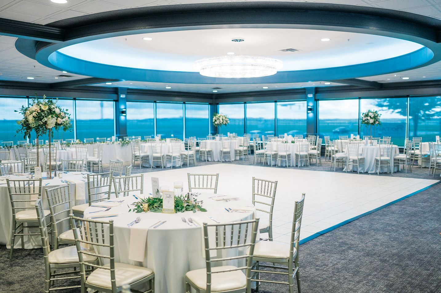 Elegant event hall with round tables, white tablecloths, and silver chairs set around a central white dance floor.