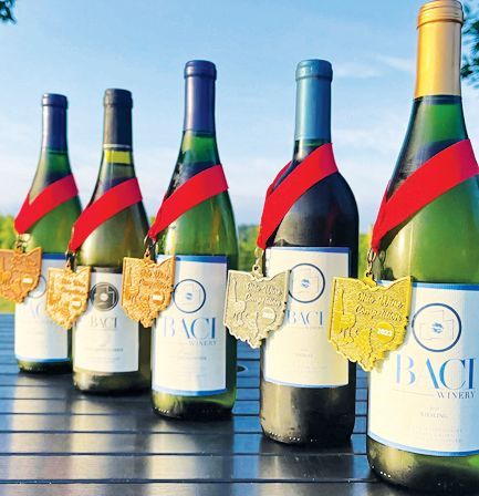 Five Baci Winery wine bottles lined up on a table, each wearing a red ribbon with an Ohio-shaped award medal.