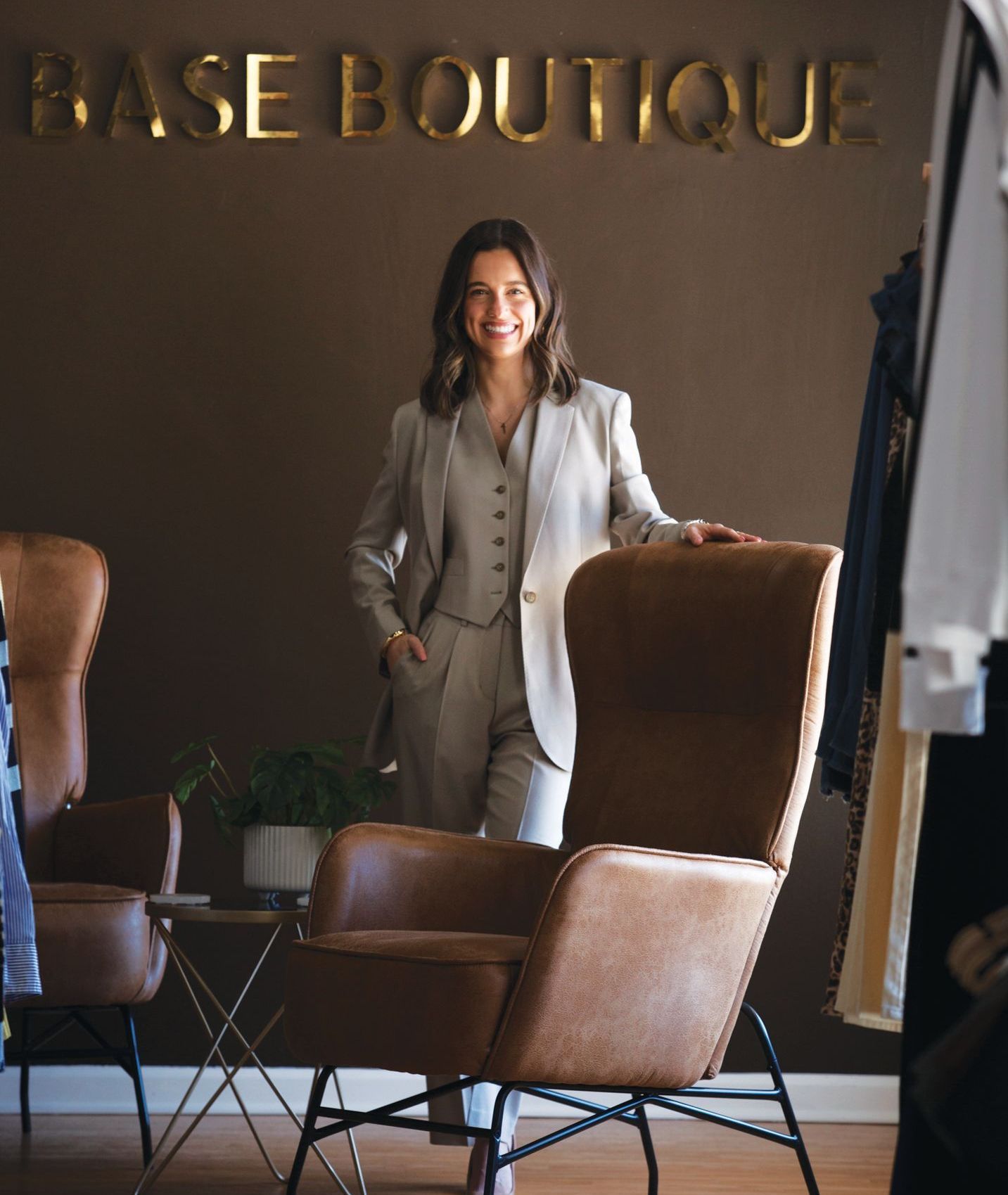 A smiling person in a light grey suit stands behind a leather armchair in front of a wall sign reading BASE BOUTIQUE.
