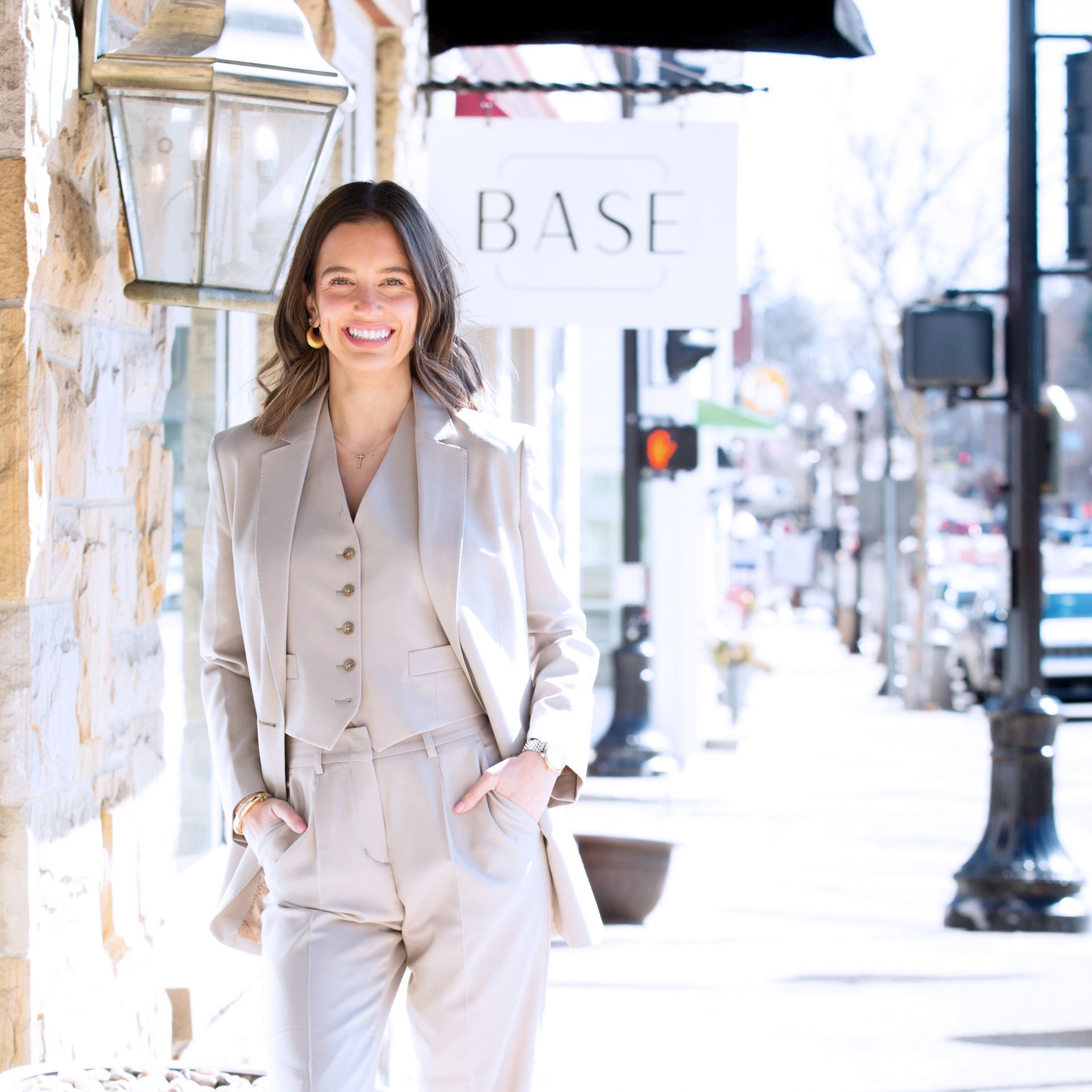 A smiling person in a beige vest and blazer suit posing on a sidewalk in front of a storefront with a