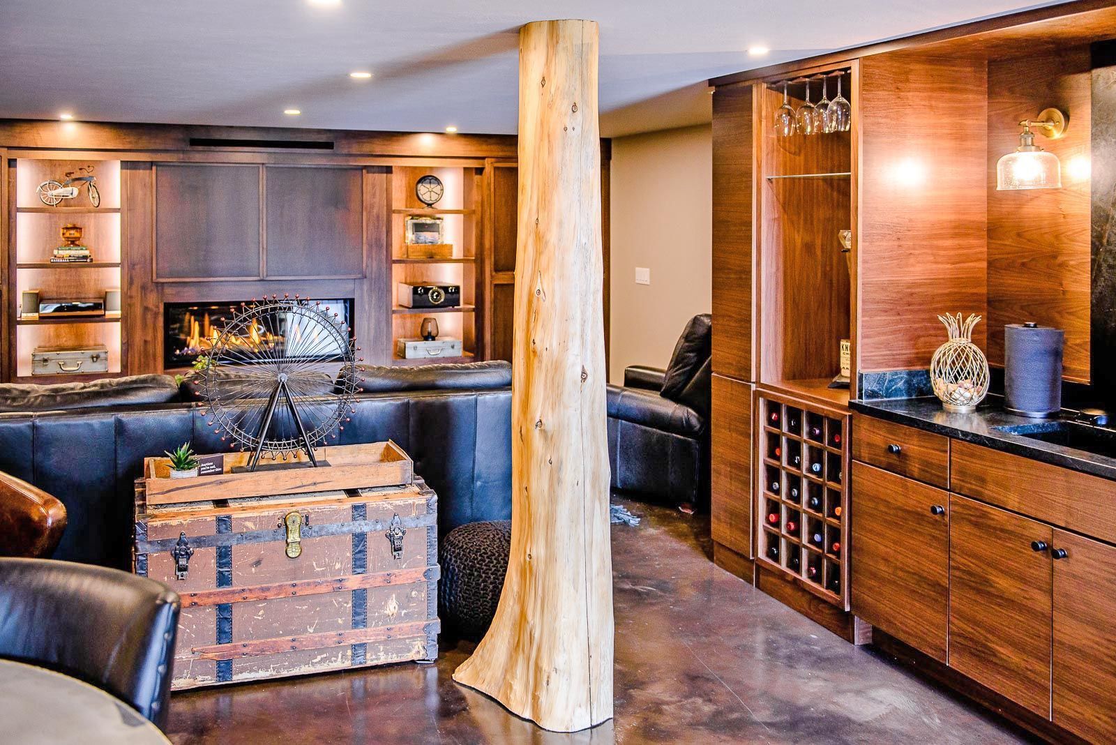 A basement living area with a leather sofa, rustic trunk, fireplace, and a bar featuring a natural wood pillar support.