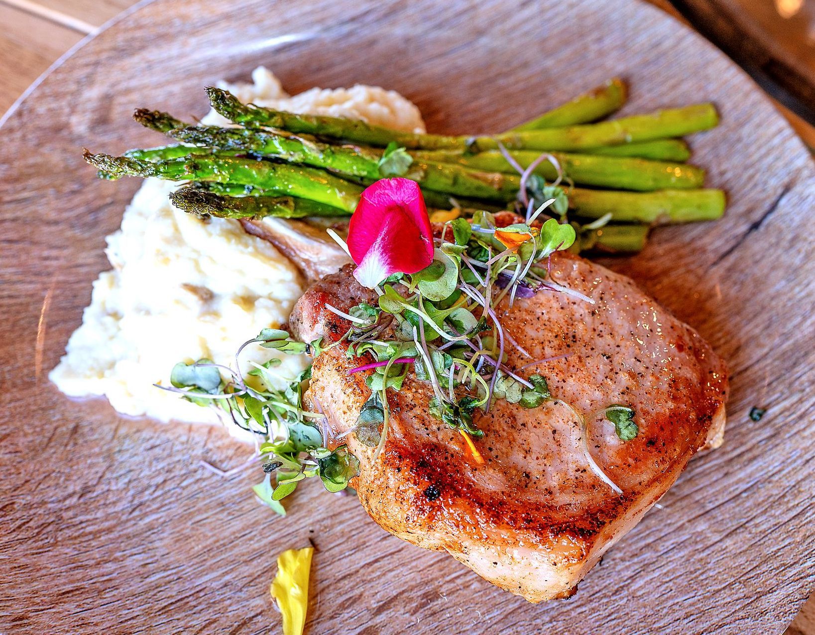 A seared pork chop with mashed potatoes and grilled asparagus, topped with fresh microgreens and an edible flower.