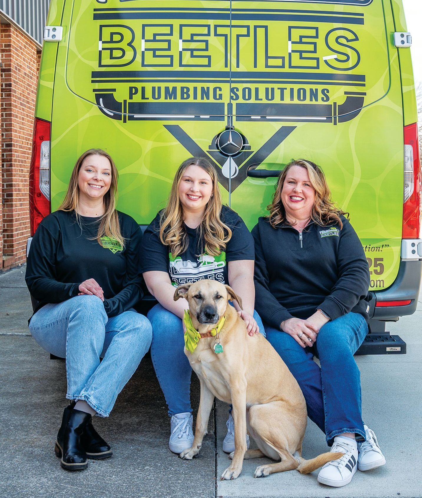 Three people sit on the ground with a tan dog in front of a bright green 