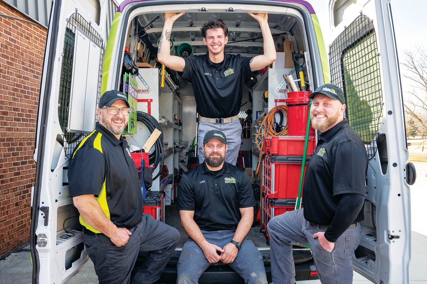 Four people in black company uniforms pose inside and around a utility van, smiling at the camera.