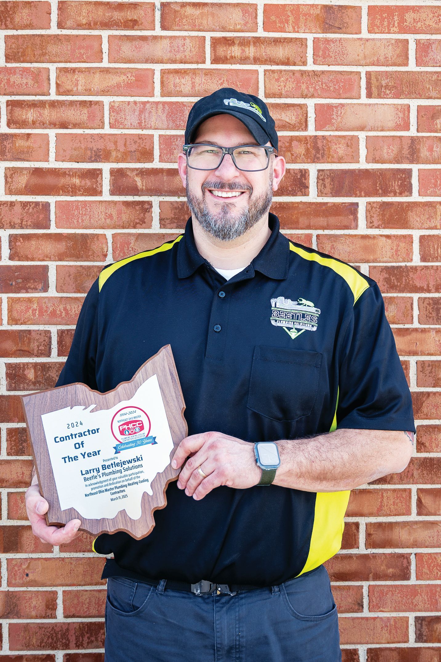 A smiling person in a black polo shirt and cap holds an Ohio-shaped award plaque against a red brick wall.
