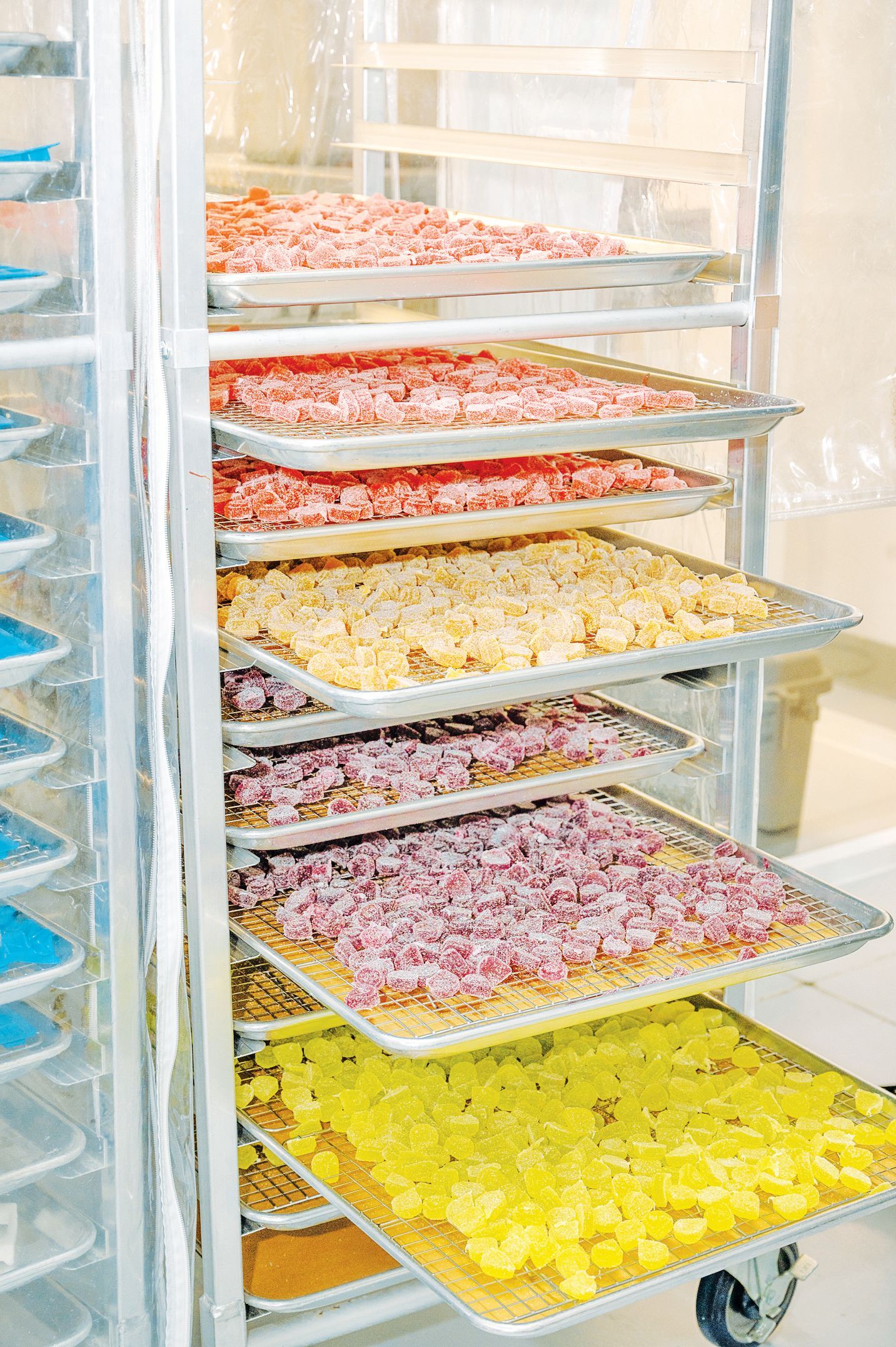 A tall metal rolling rack holds multiple sheet pans filled with colorful food, including red, yellow, and textured items.
