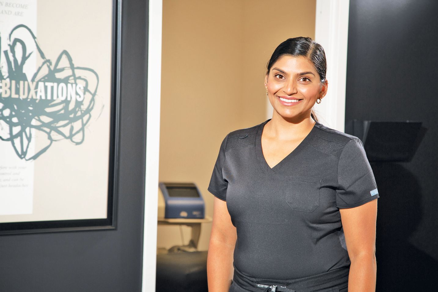 A smiling person in a dark grey V-neck scrub top stands in front of a white wall next to a framed sign.