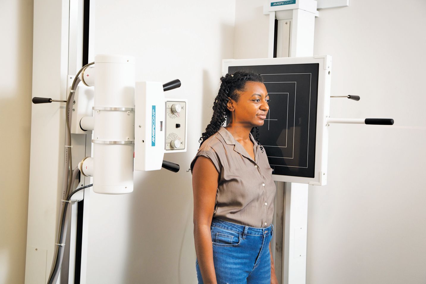 A person stands in a clinic, positioned against a vertical digital X-ray detector for imaging.