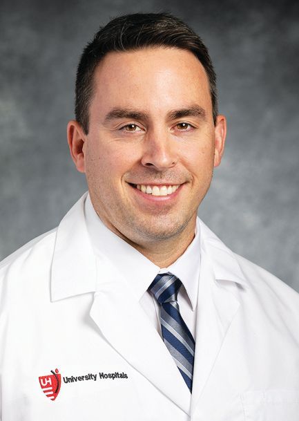 A smiling medical professional wearing a white coat with a University Hospitals logo over a blue striped tie.