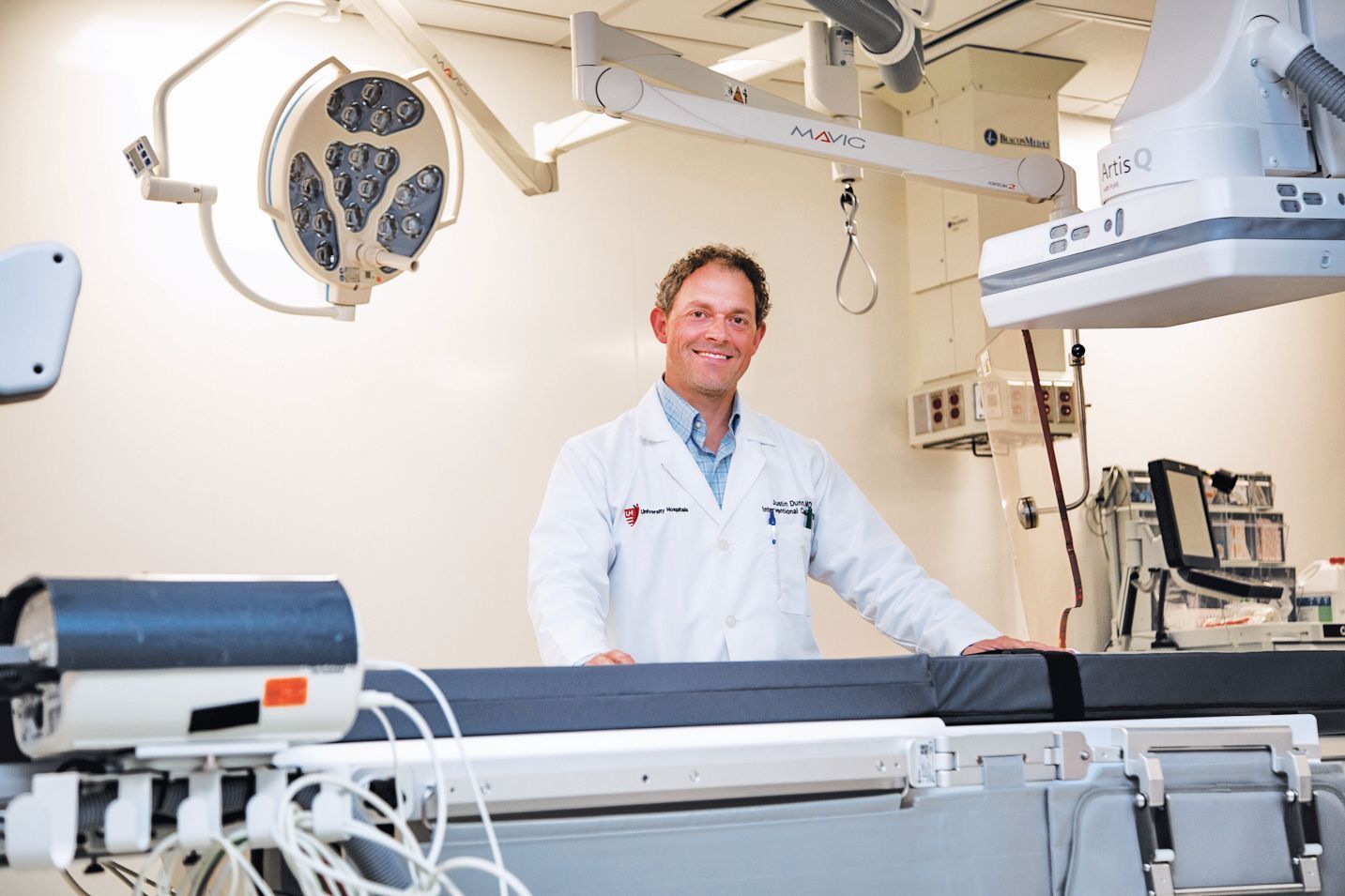 A smiling medical professional in a white coat stands in a sterile operating room with surgical equipment.