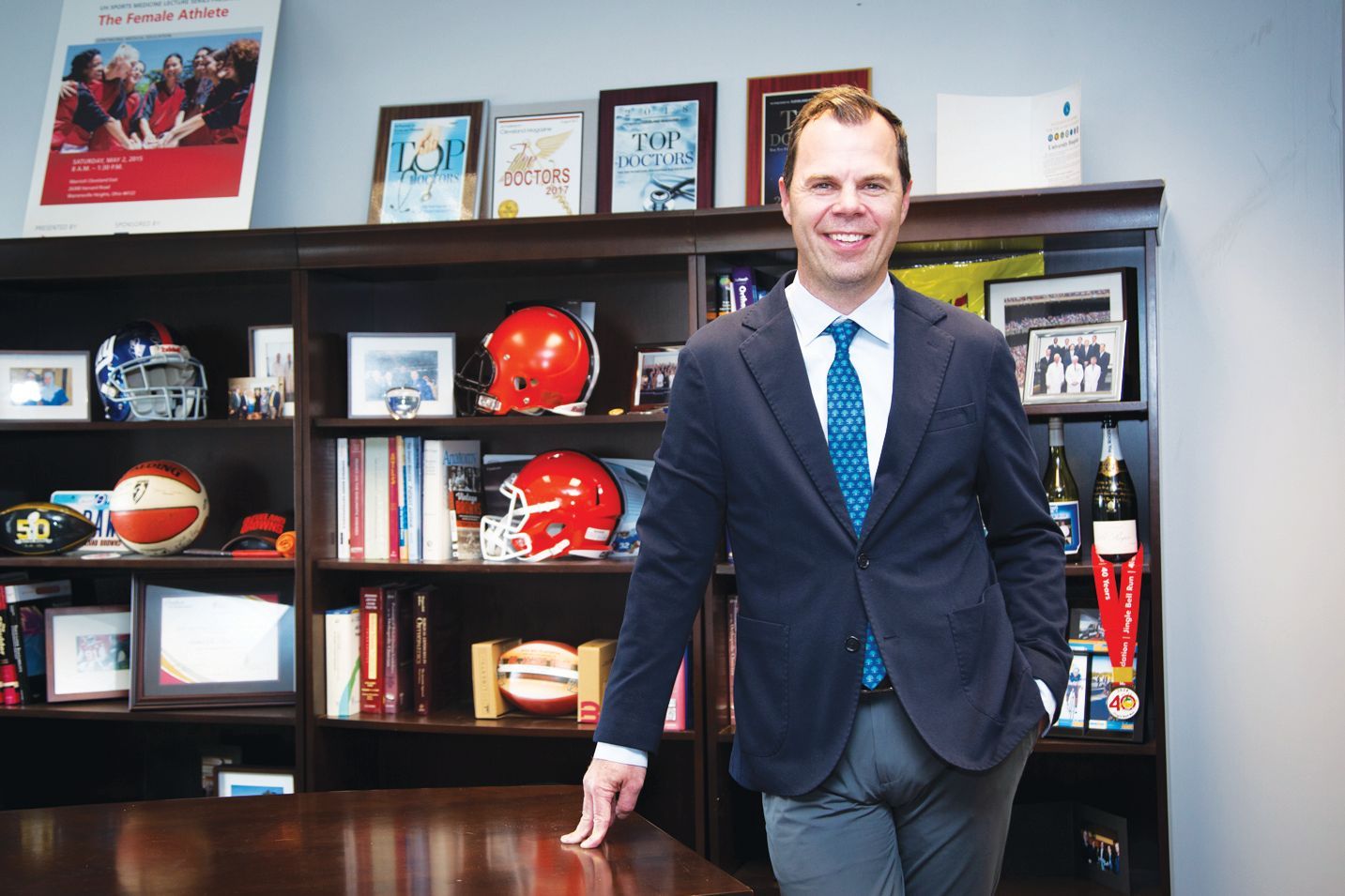 A smiling person in a suit and blue patterned tie leans against a dark wooden bookshelf filled with sports memorabilia.