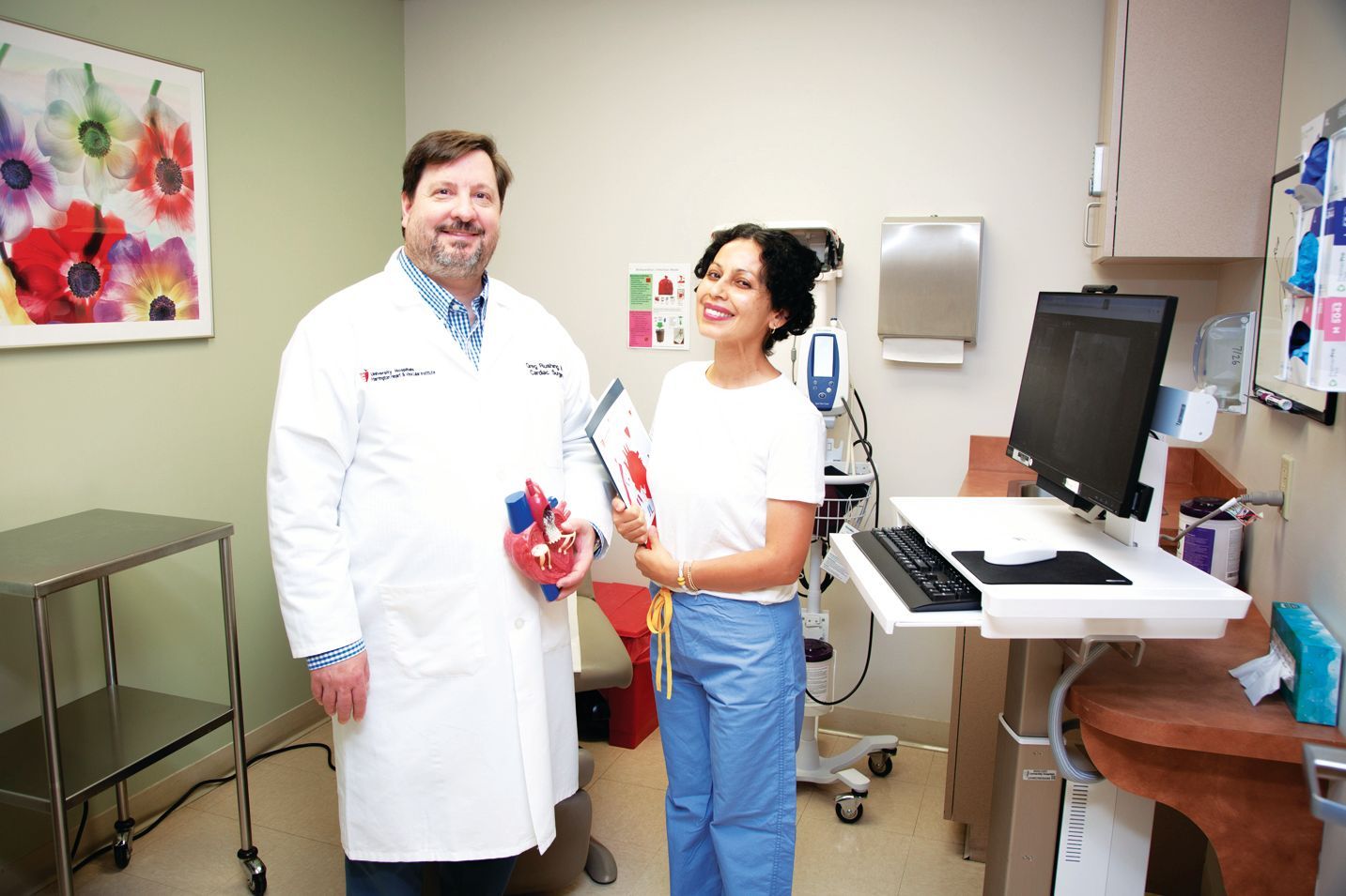 A doctor holding a heart model stands next to a person holding a folder in a medical office with a computer station.