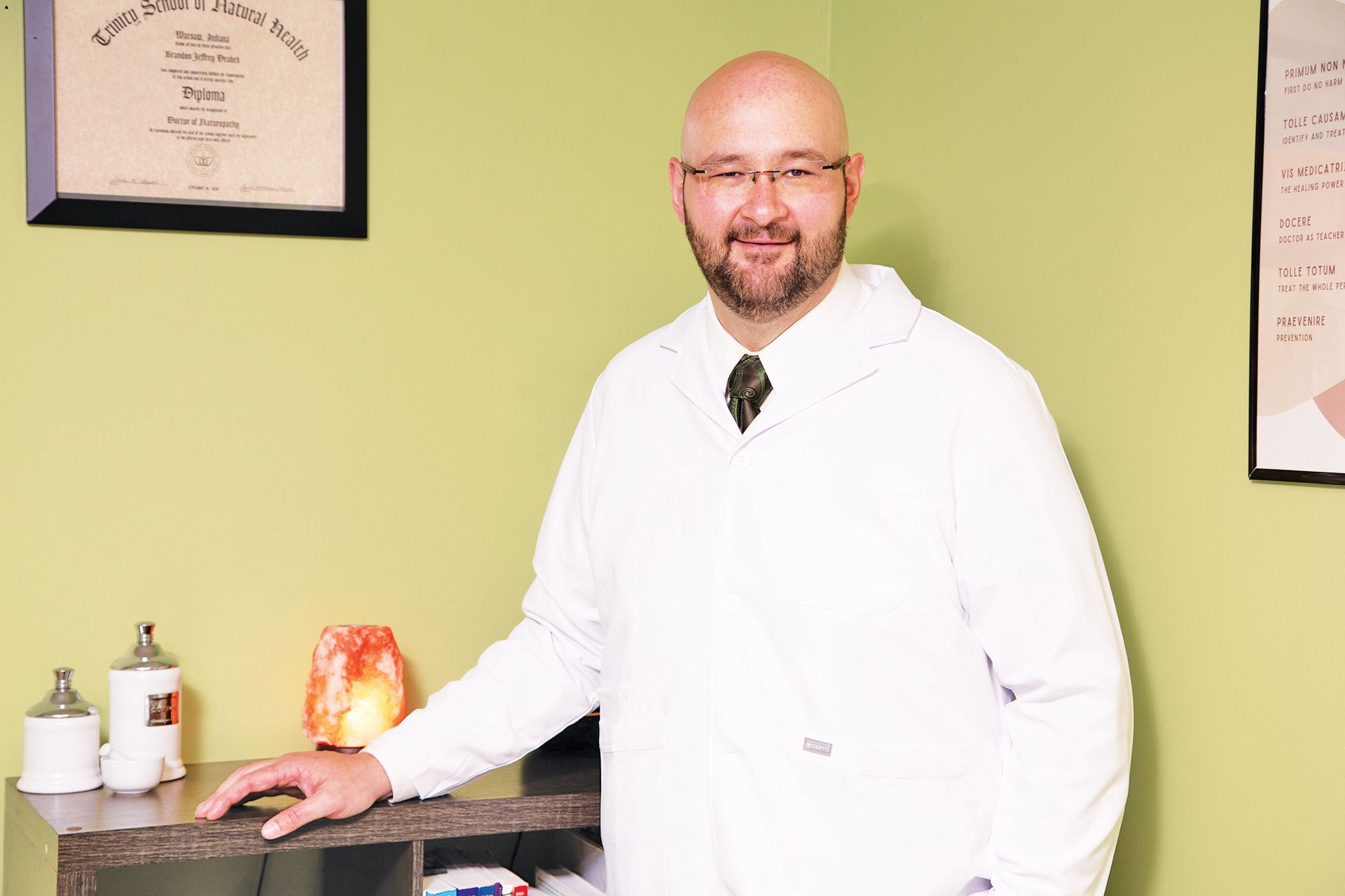 A smiling person wearing glasses and a white lab coat stands in an office with a diploma and a salt lamp on the shelves.