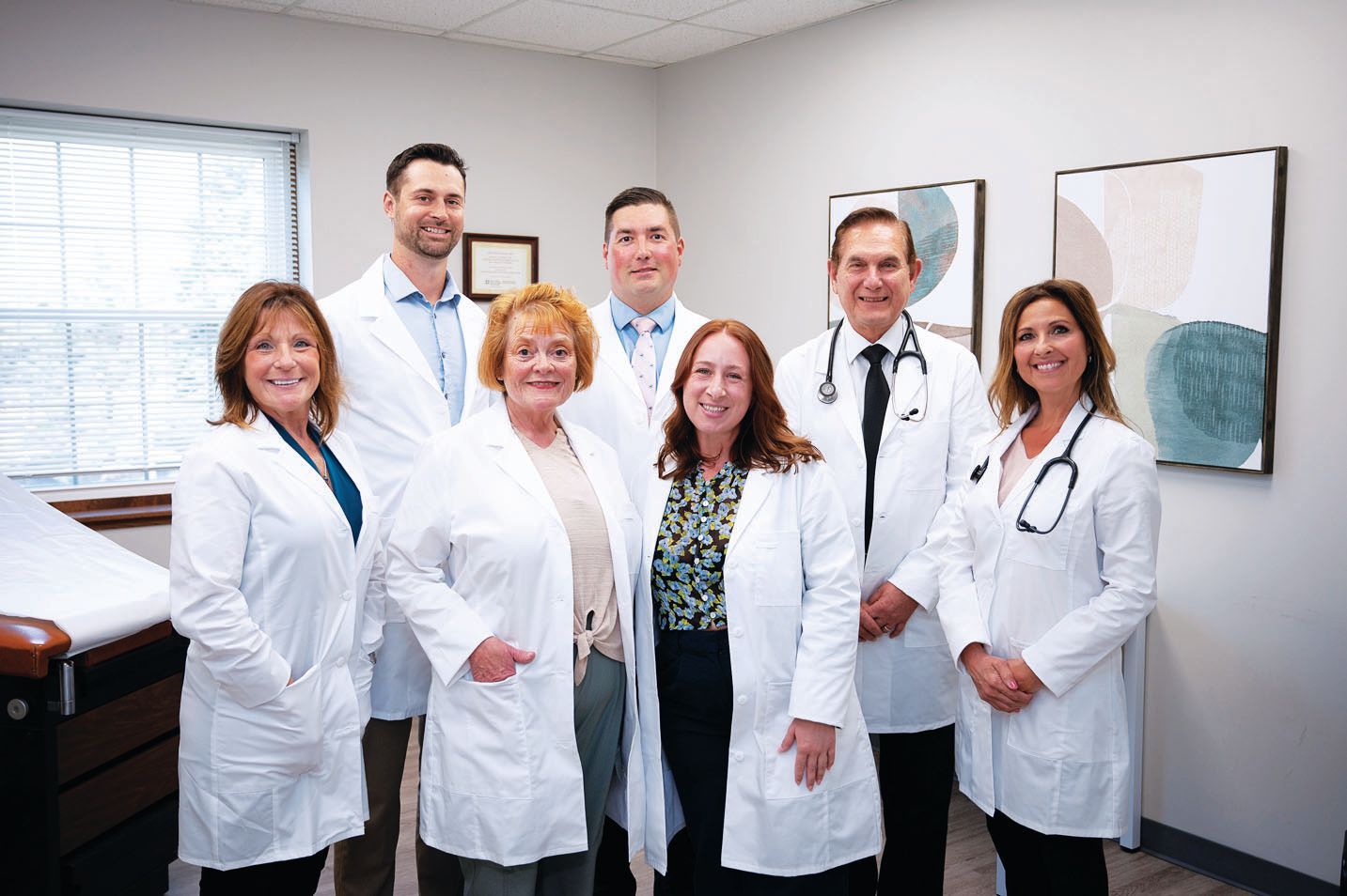 Seven medical professionals in white coats posing for a group portrait in a brightly lit doctor's office.