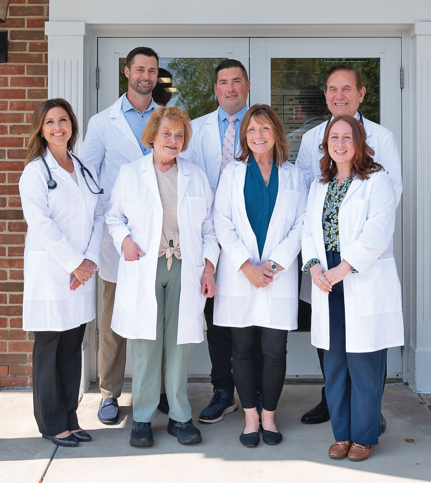 A group of seven medical professionals in white coats posing together in front of a building entrance.