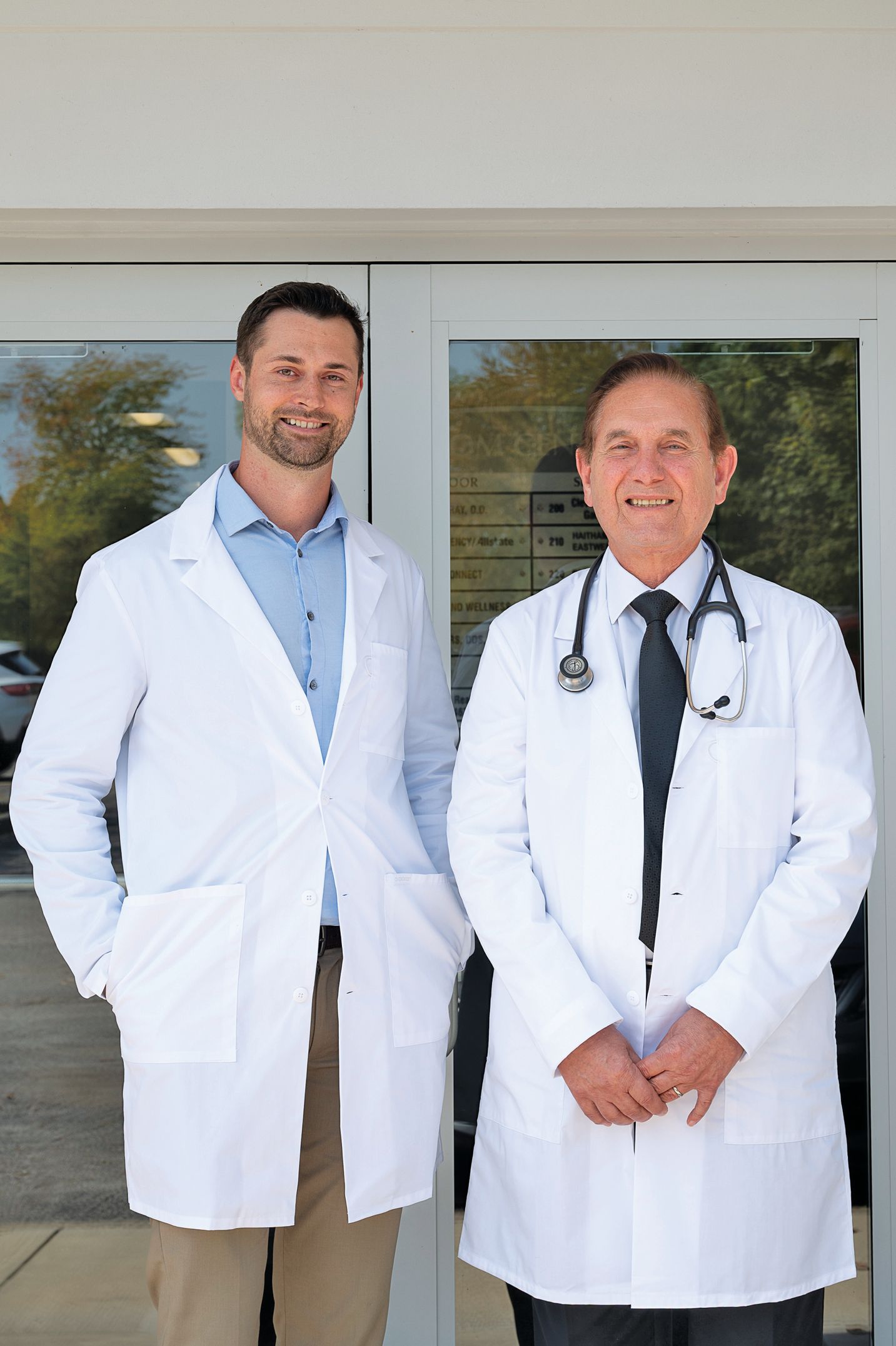 Two medical professionals in white lab coats standing together in front of a glass door entrance.
