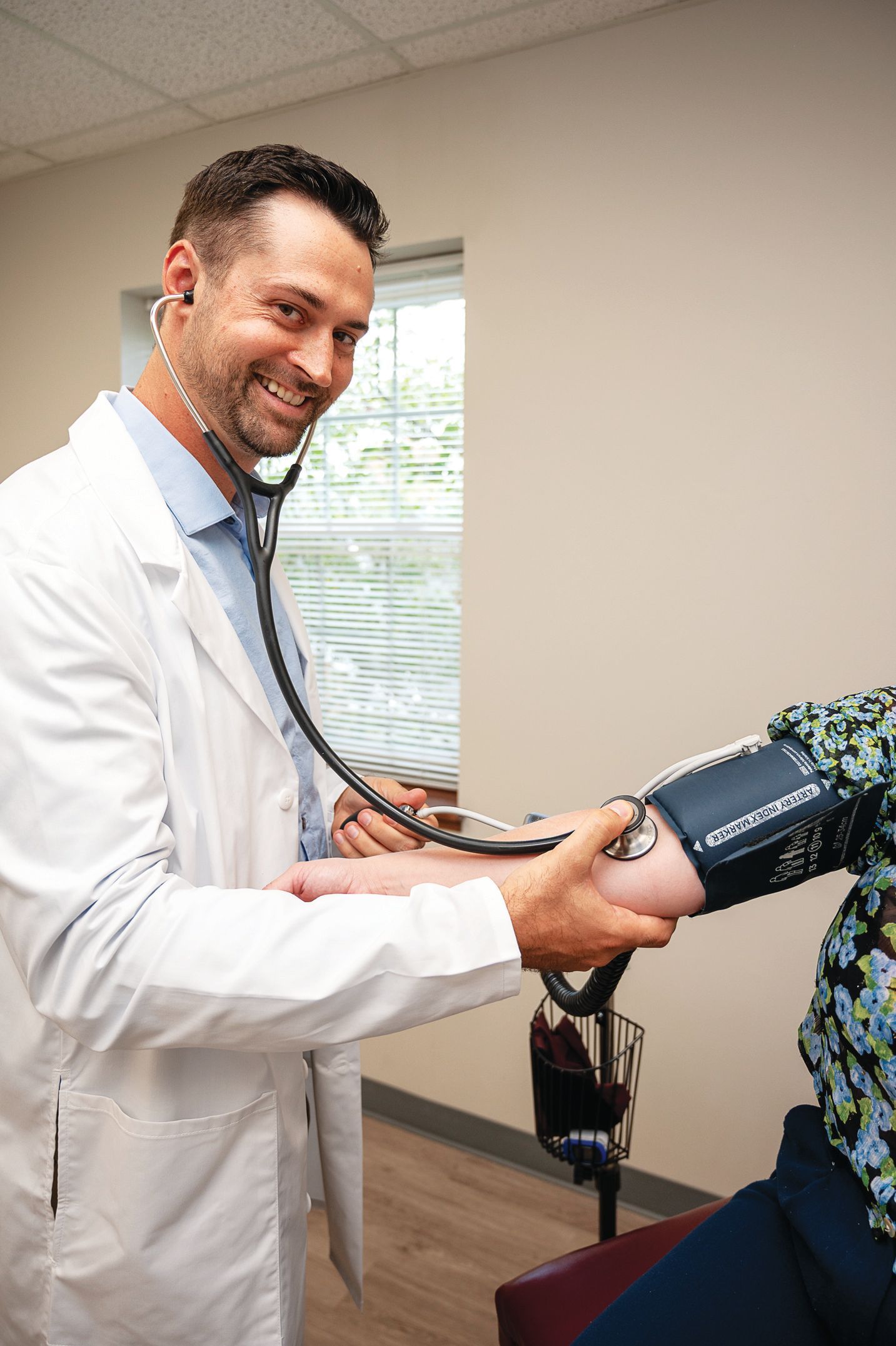 A smiling healthcare professional uses a stethoscope to measure a patient’s blood pressure in an office setting.