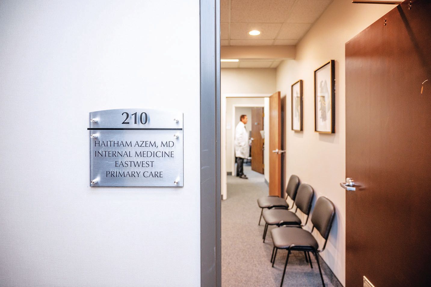 A medical office hallway at room 210, with chairs against the wall and a healthcare professional in the background.