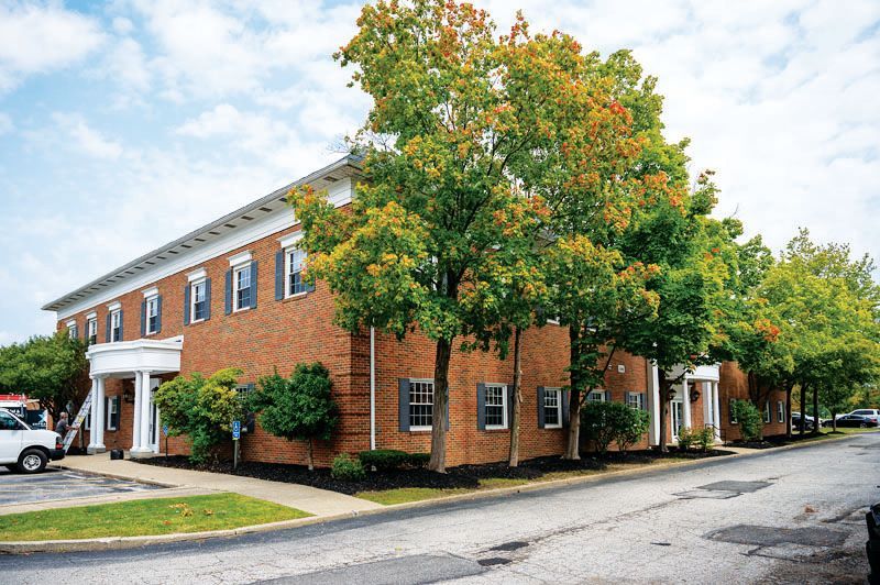 A brick, two-story commercial building with white columns and green trees on a paved lot under a cloudy sky.