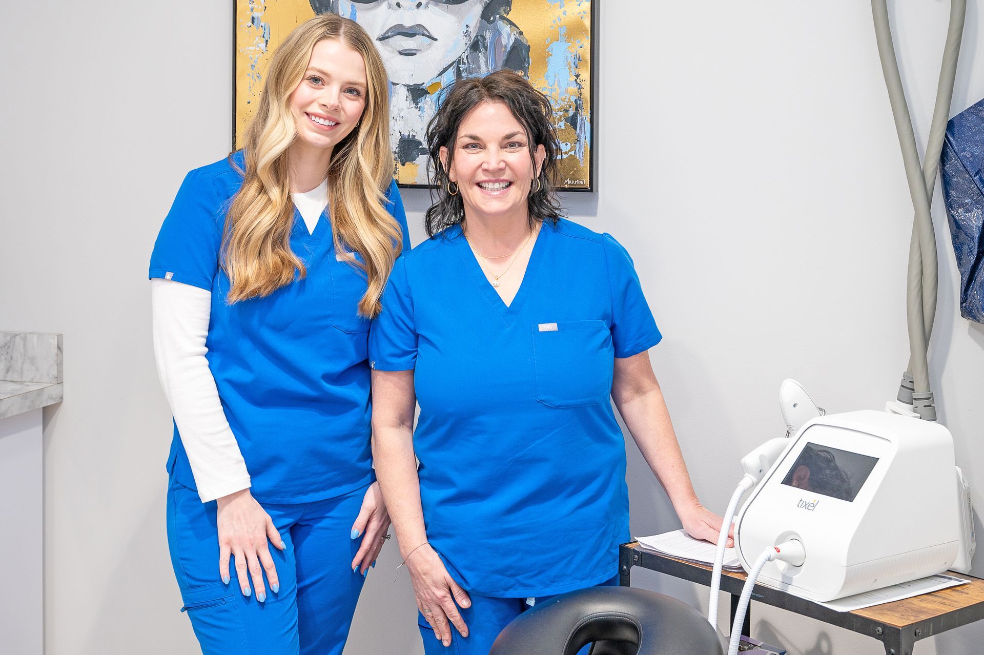 Two people in blue medical scrubs stand in a treatment room next to medical equipment and artwork.