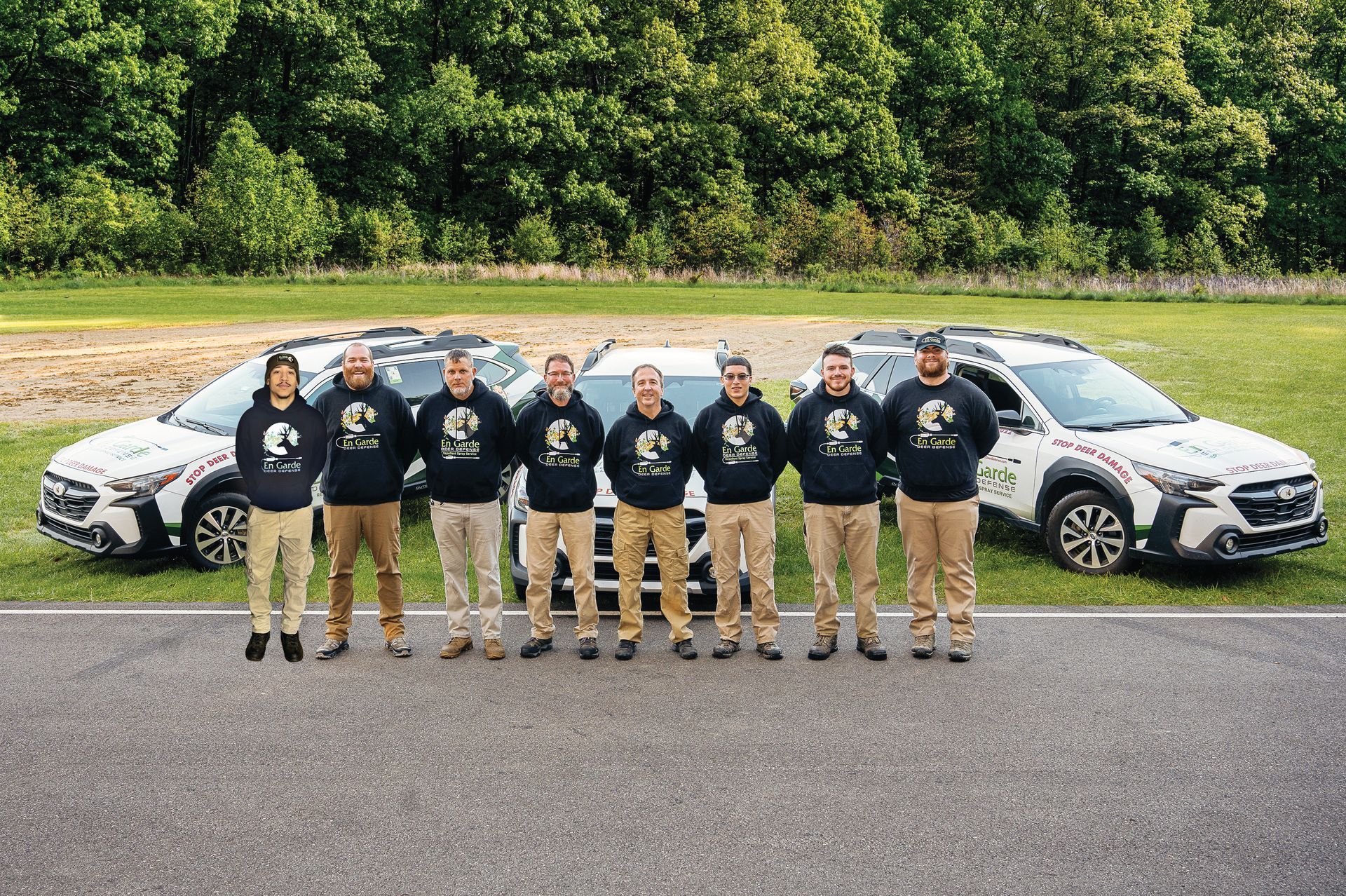 Eight people wearing matching dark sweatshirts stand in a line outdoors between two white vehicles.