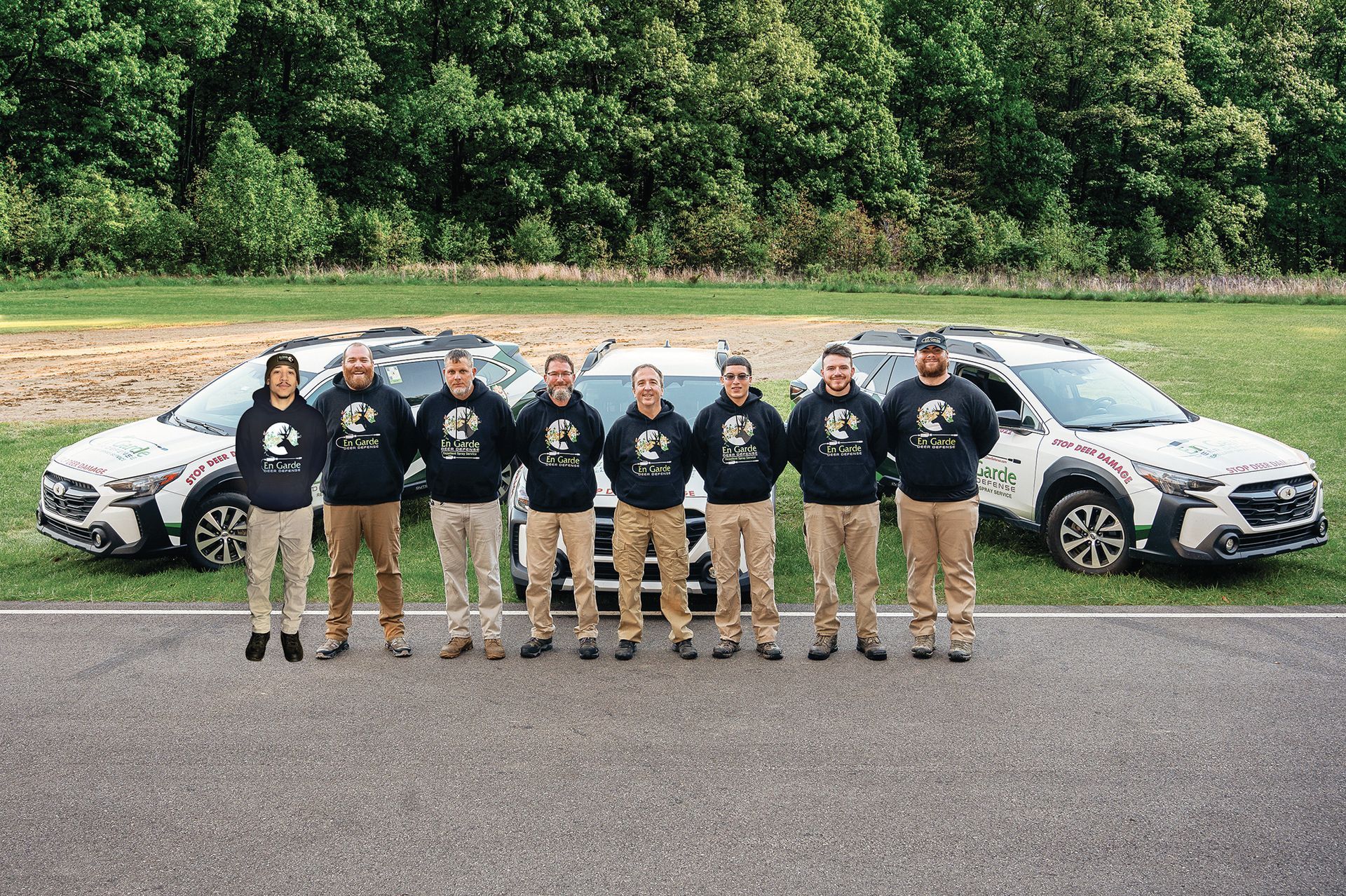 Eight people in matching navy hoodies stand in a row before two marked patrol vehicles in a grassy outdoor setting.