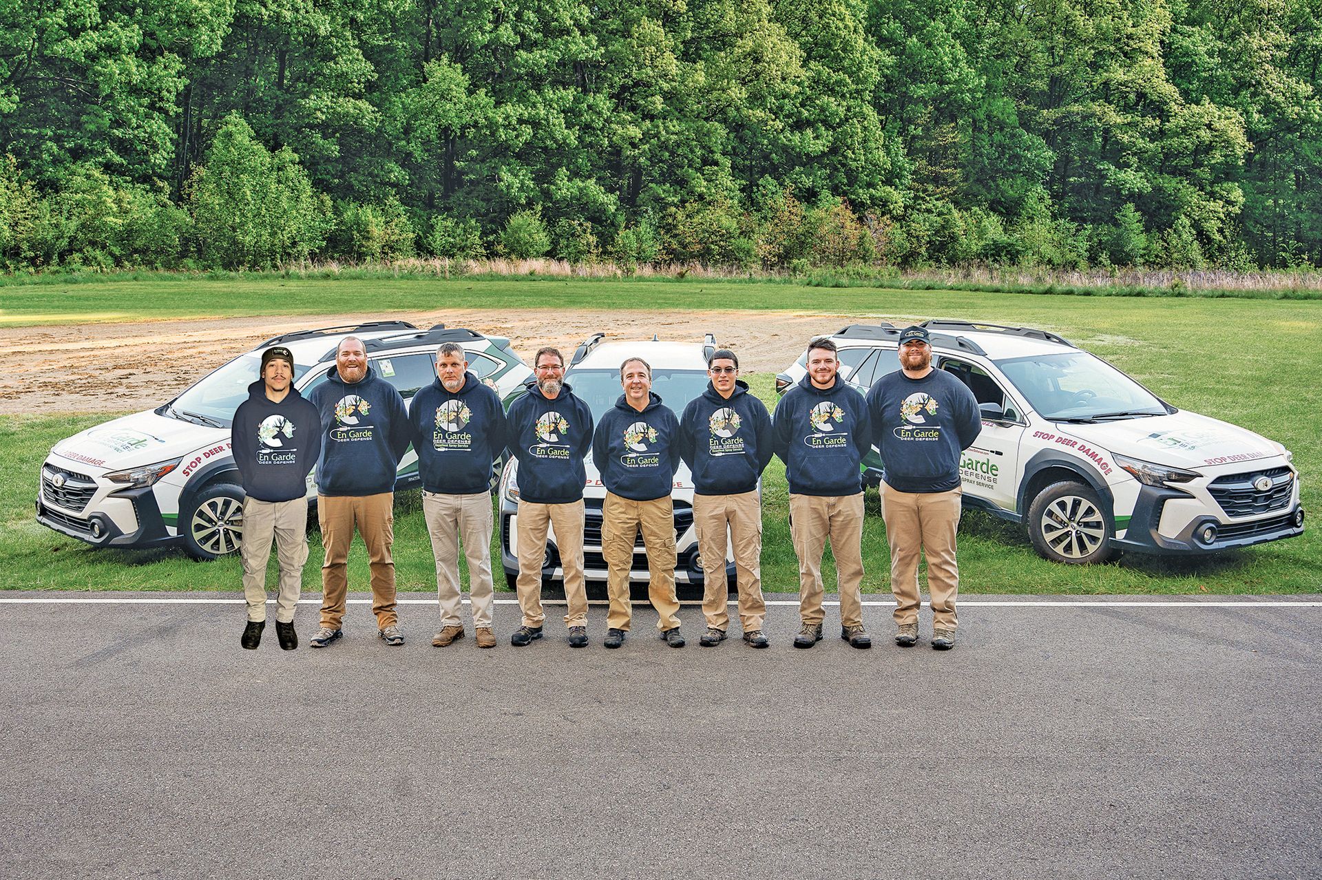 Eight people in matching navy hoodies stand in a line outdoors in front of two white SUVs on a paved lot near a grassy field.