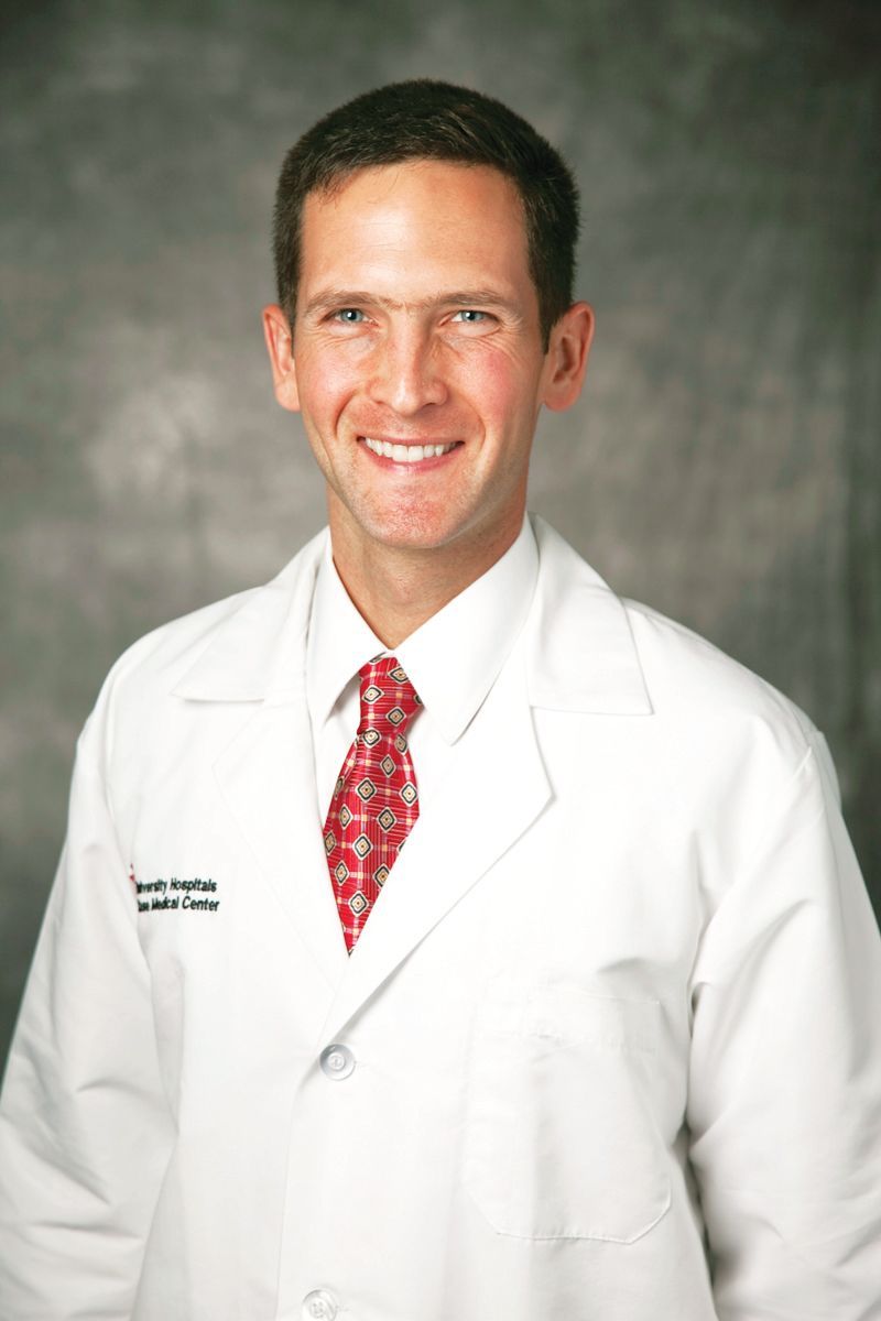 A smiling medical professional wearing a white lab coat and a red patterned tie against a gray backdrop.